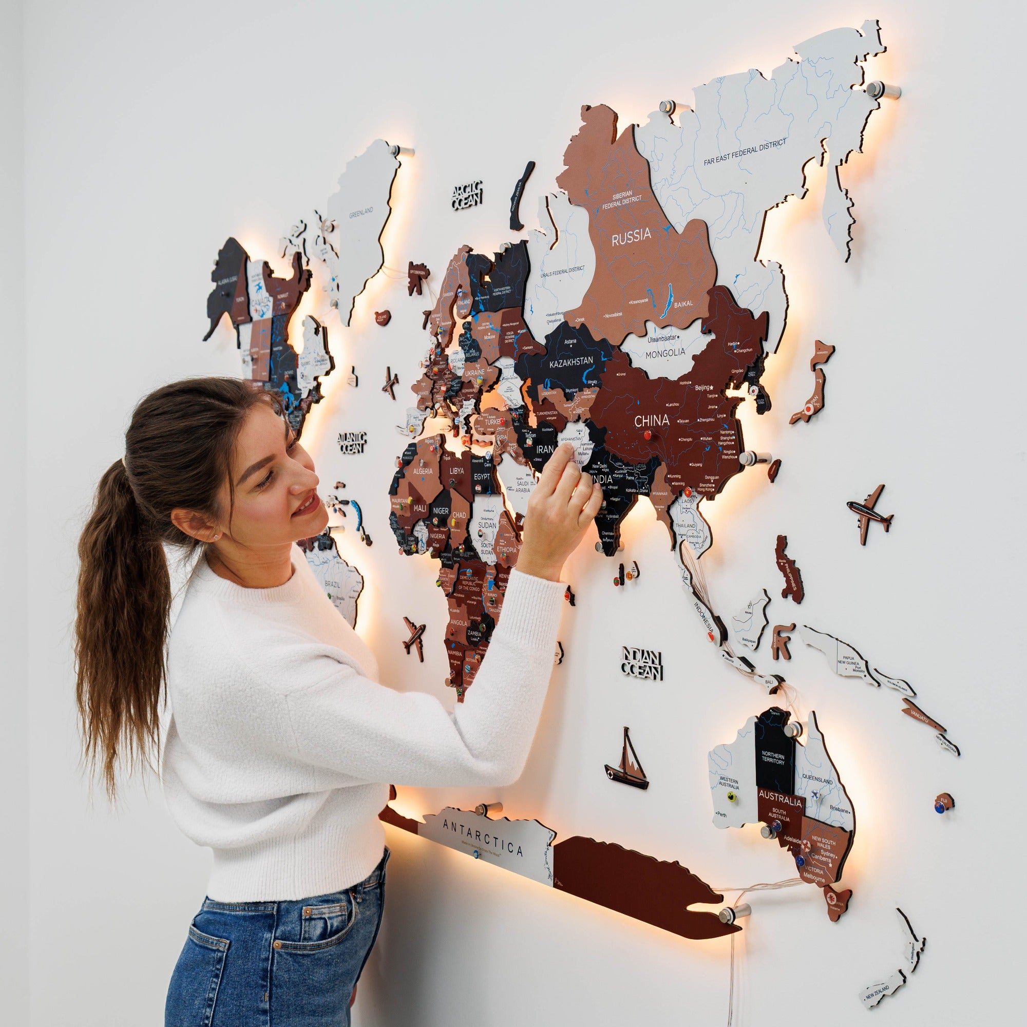 Woman taking a photo of a wooden map of the world with lights on a white wall.