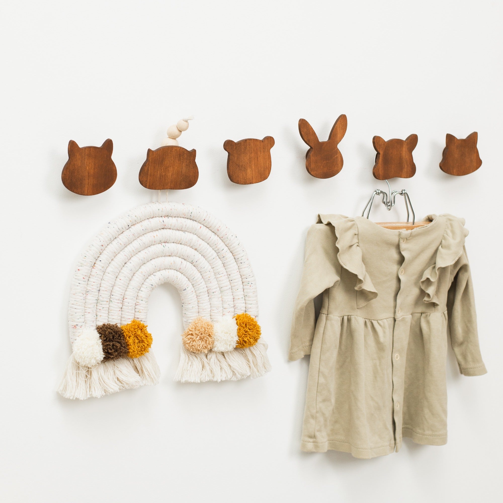 Wooden wall hooks shaped like animal ears, a macrame rainbow hanging, and a beige dress on a white background.