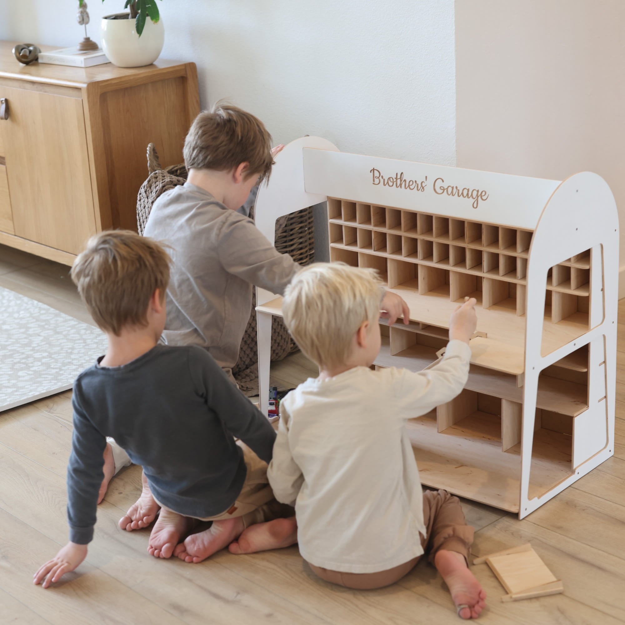 Three children sitting on a wooden floor in front of a white wooden toy organizer labeled 'Brothers' Garage' in a bright indoor room.
