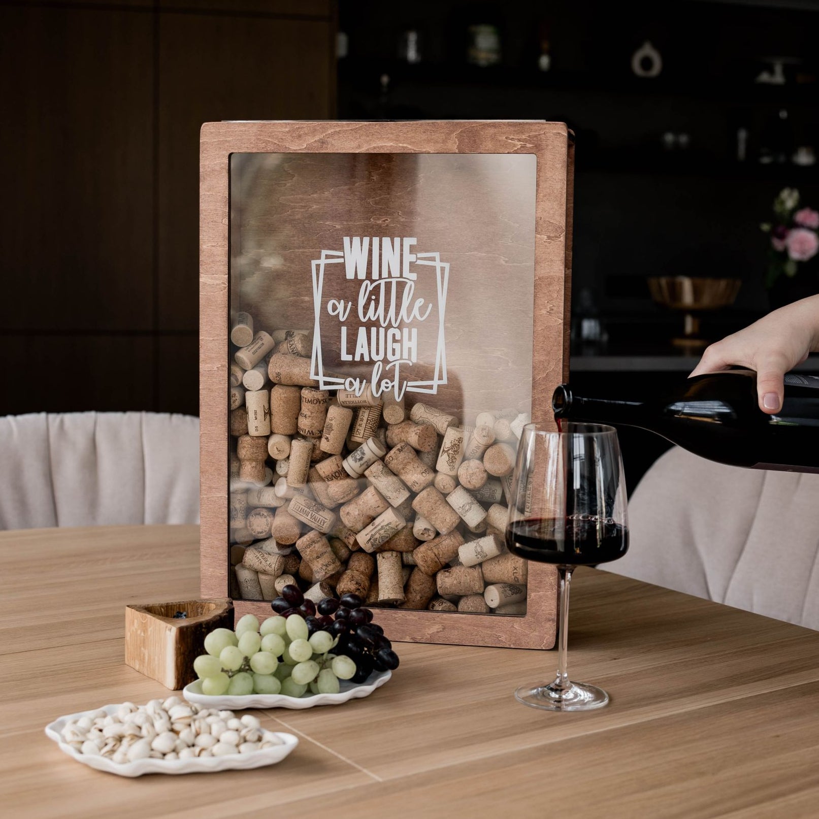 Decorative cork collector box with "Wine a little, laugh a lot" text, shown on a dining table while someone pours red wine into a glass.