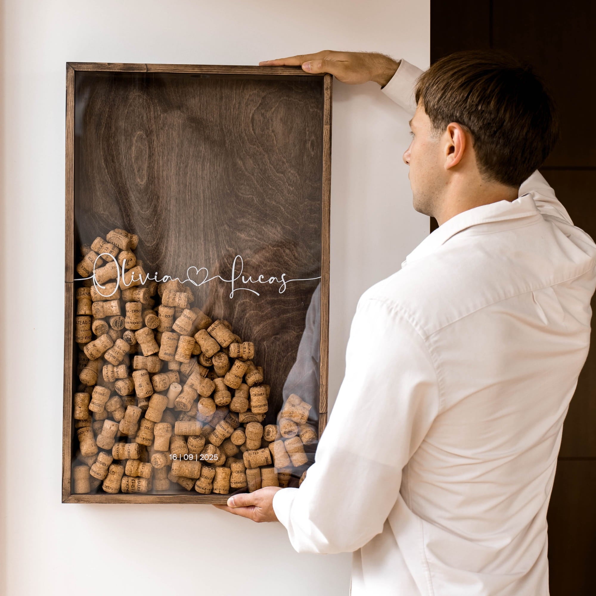 A man in a white shirt holds a framed wall display made of many wine corks on a dark wood background, with the names 'Olivia' and 'Lucas' written across the front.