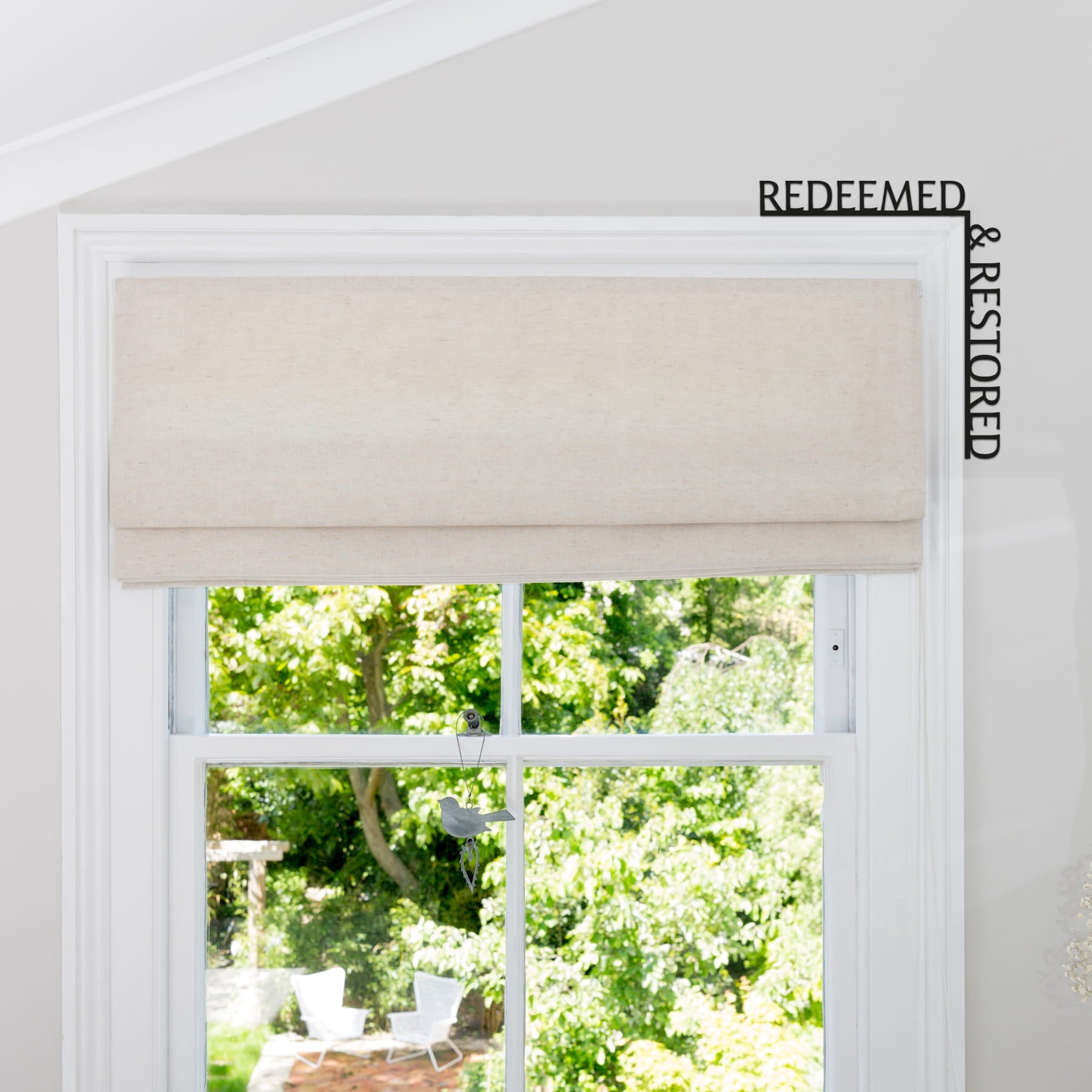 Window with beige roman blind and view of greenery outside, featuring 'REDEEMED & RESTORED' text.