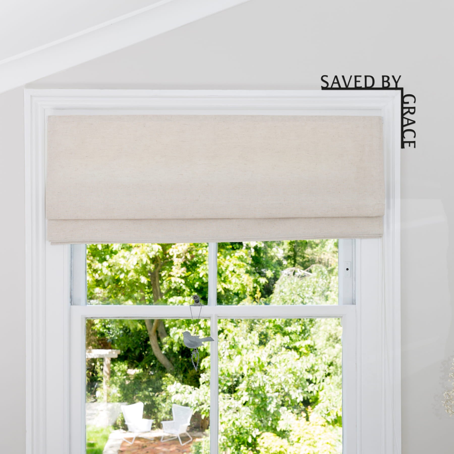 Window with beige roman blind and view of greenery outside, featuring 'Saved by Grace' branding.