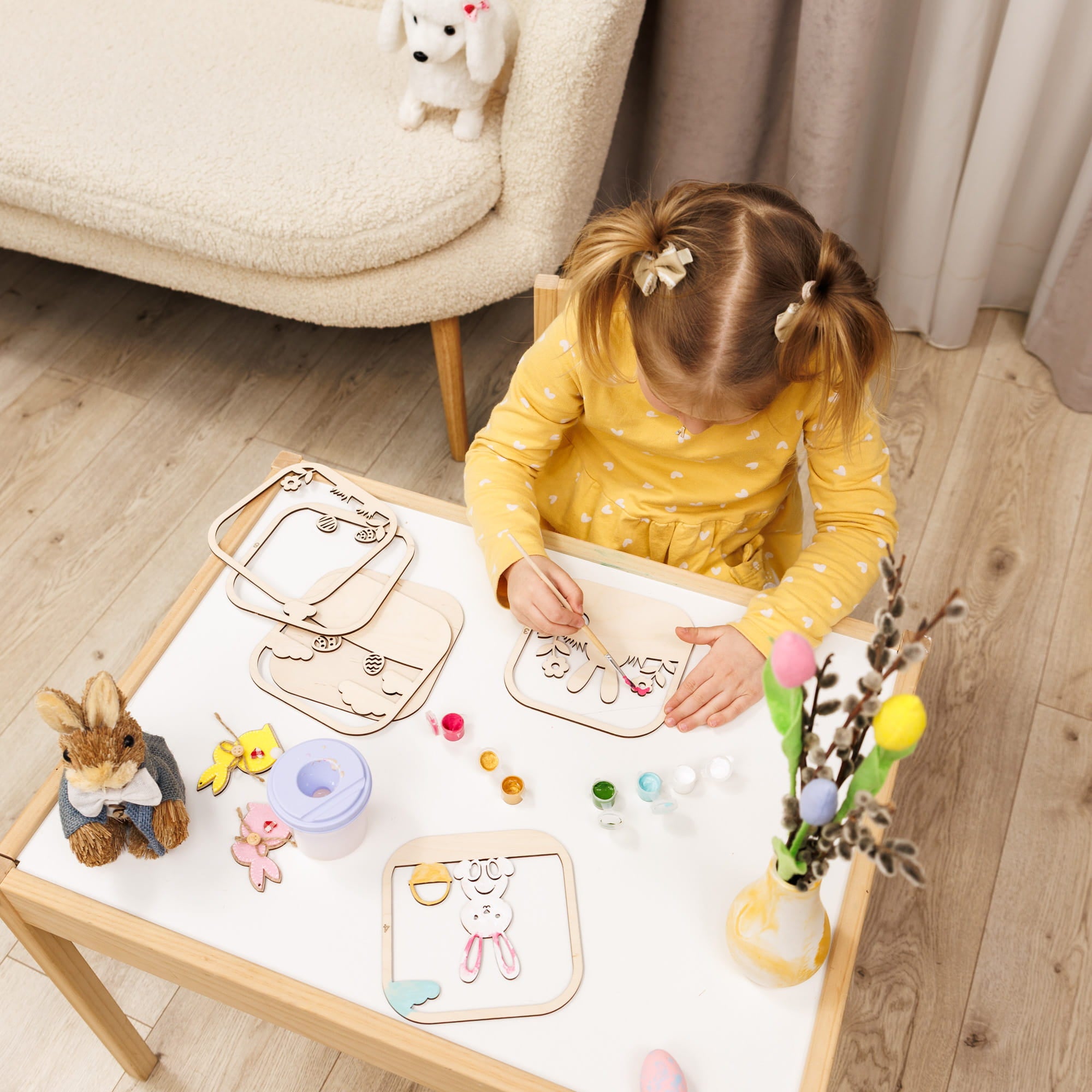 Child in a yellow polka dot dress sitting at a small table with art supplies, drawing on paper.