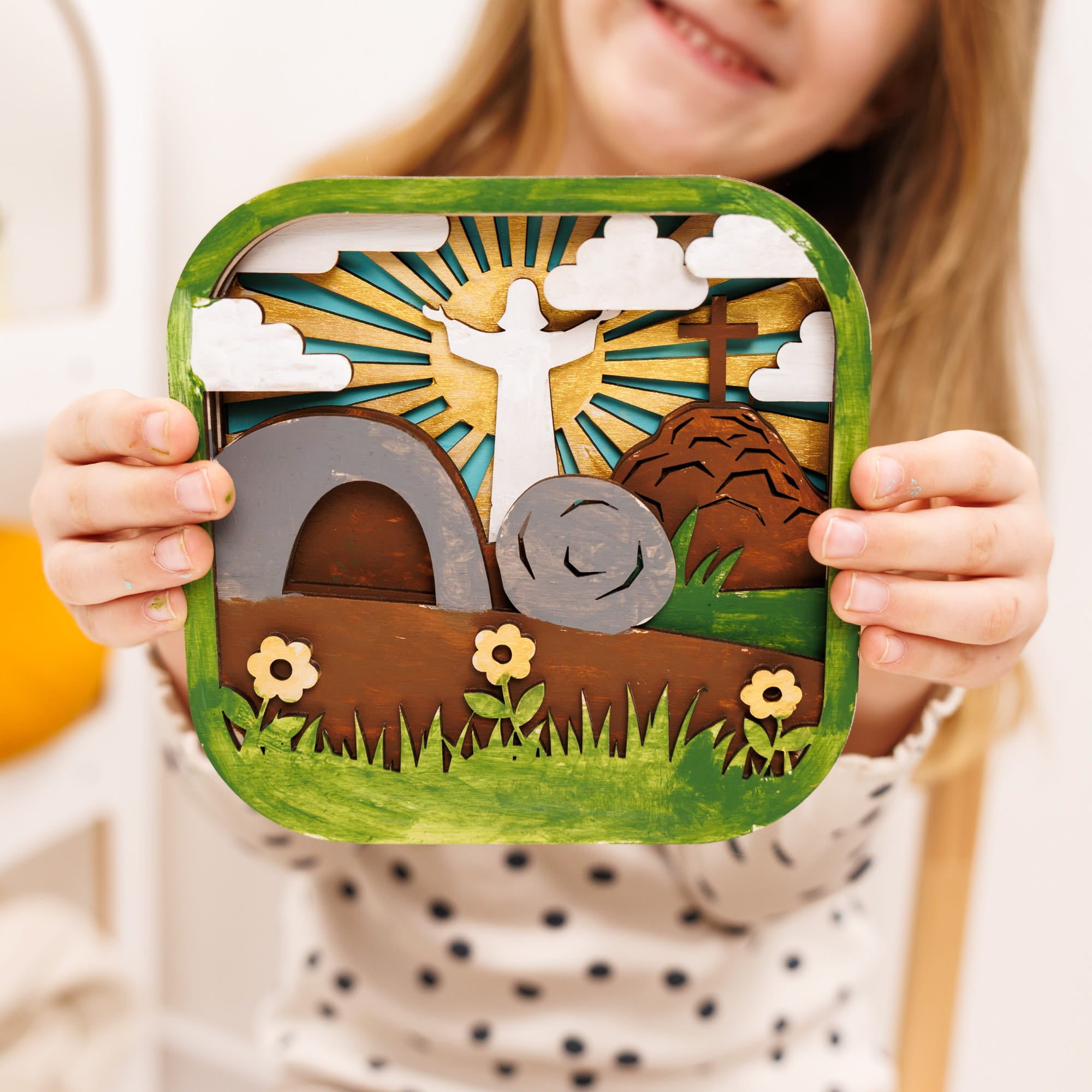 Child holding a religious-themed wooden cutout with a cross, sun, and other elements.