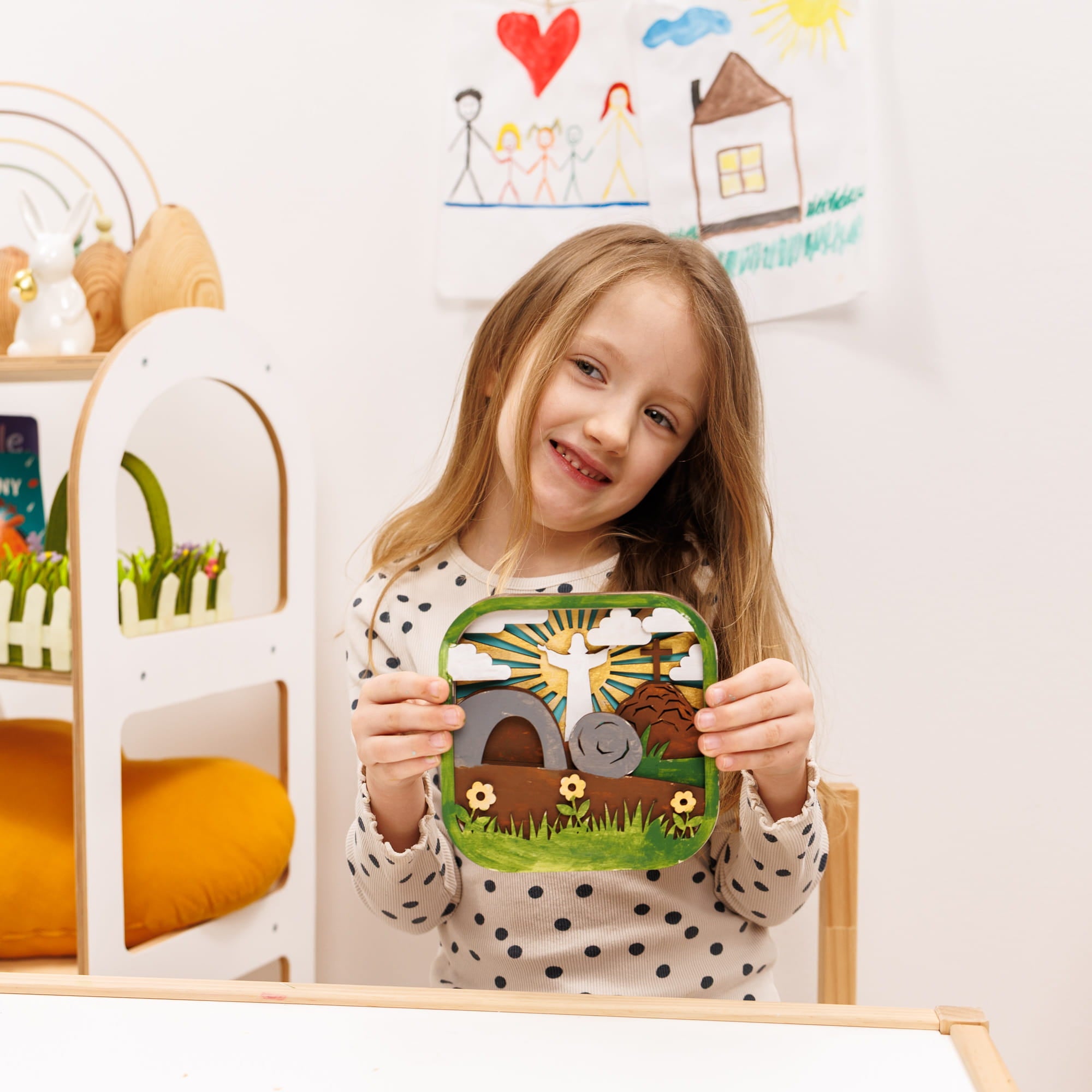 Young girl holding a felt craft with a house design in a playroom setting.