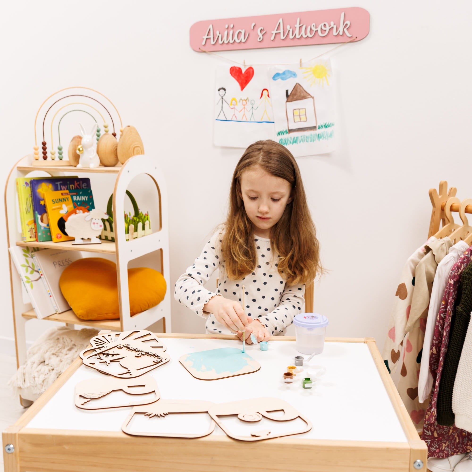 Child at a table with art supplies and a shelf in a room labeled 'Aria's Artwork'.