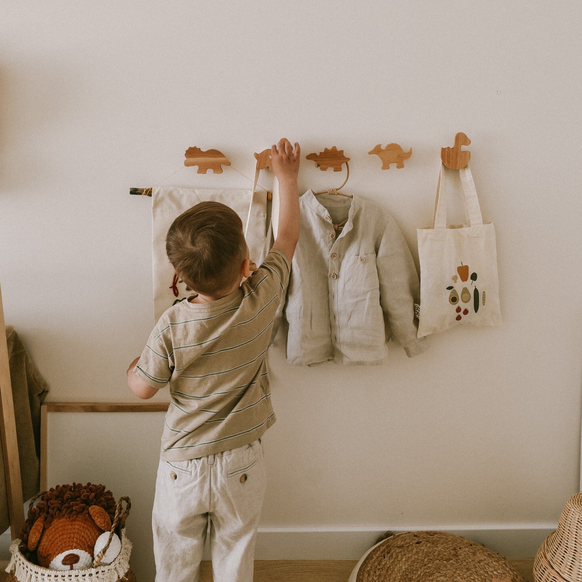 Child hanging a shirt on a hook in a room with a basket of toys and a bag.