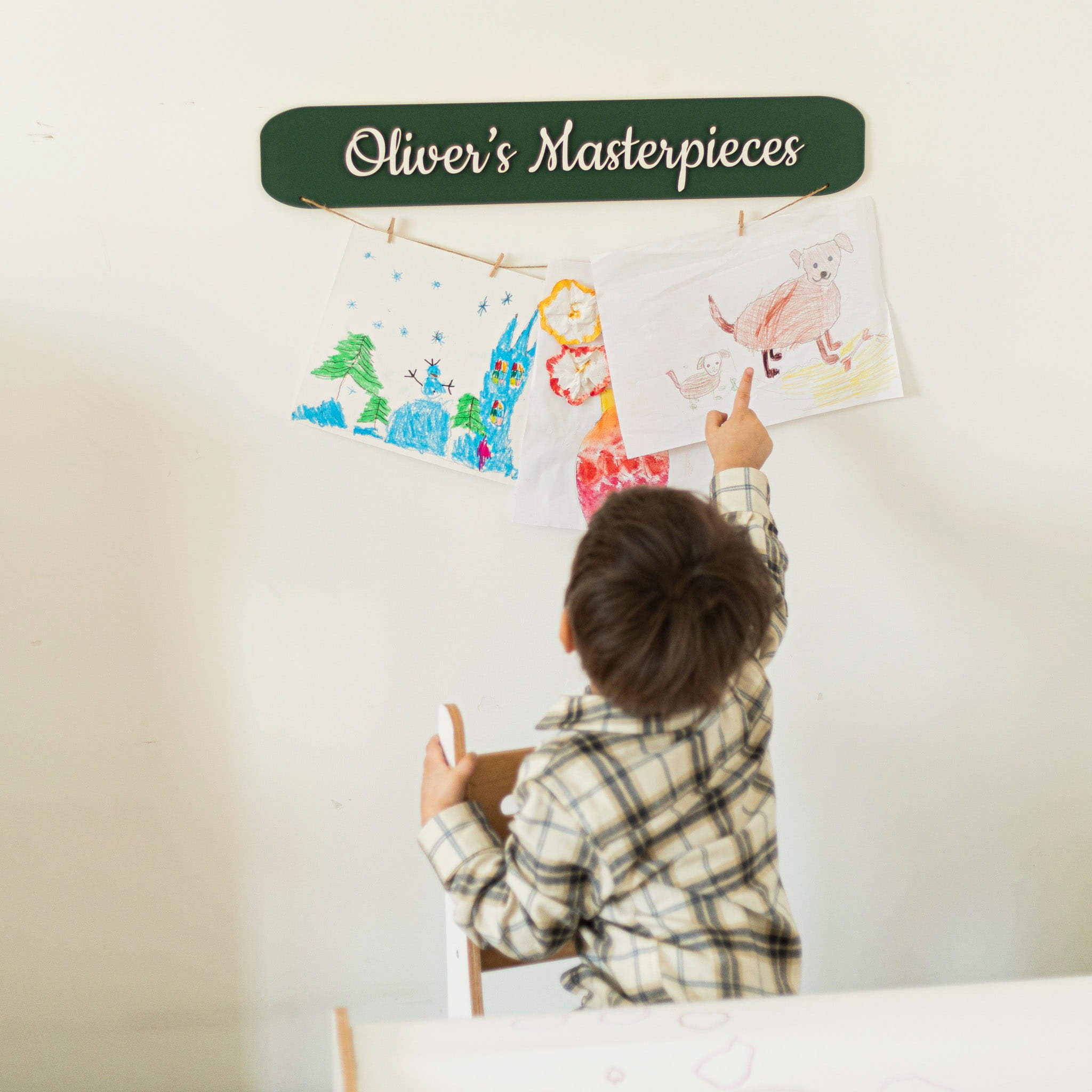 Child hanging drawings on a wall with a sign that reads 'Oliver's Masterpieces'.