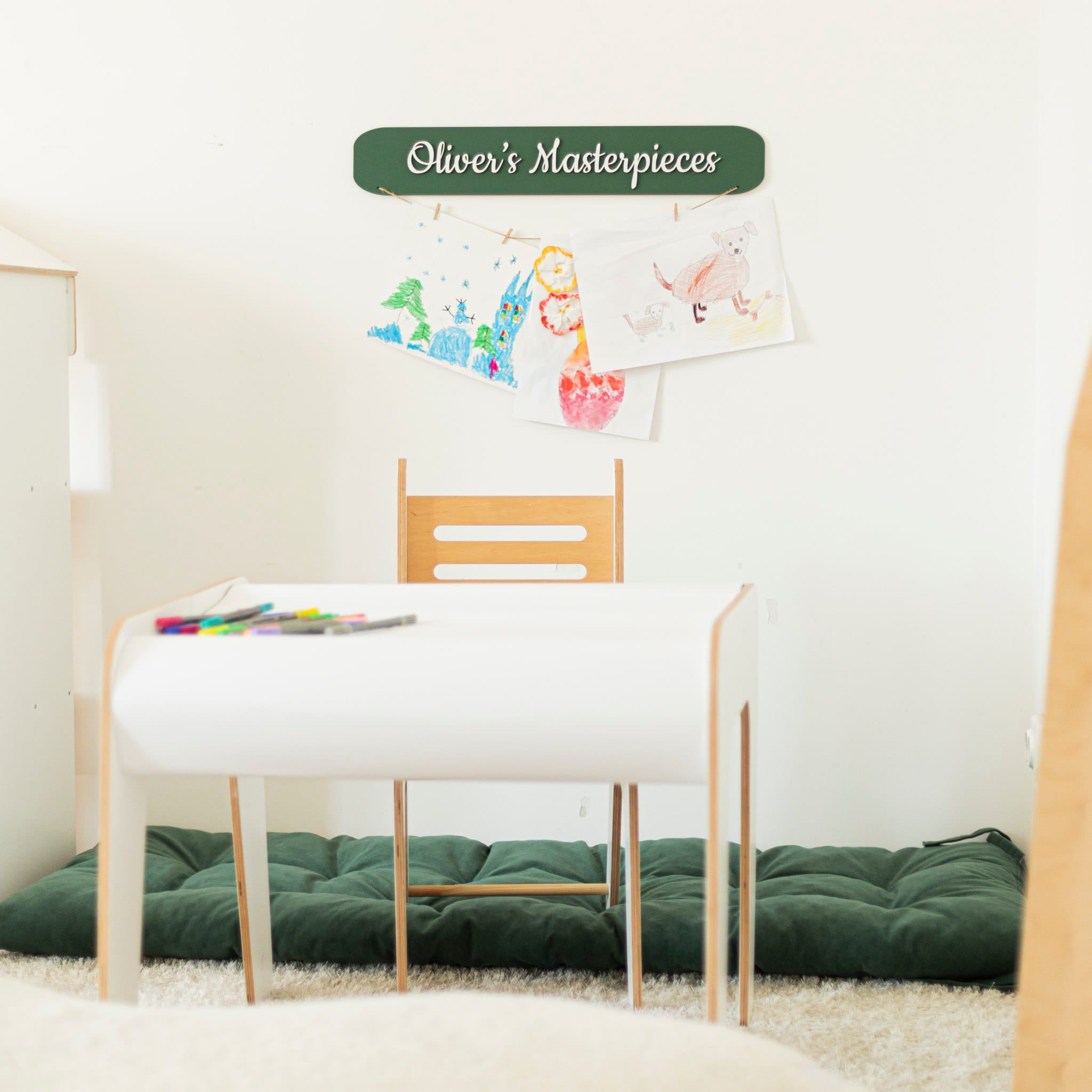 Children's drawing on a wall above a white table with a chair, labeled 'Oliver's Masterpieces'.