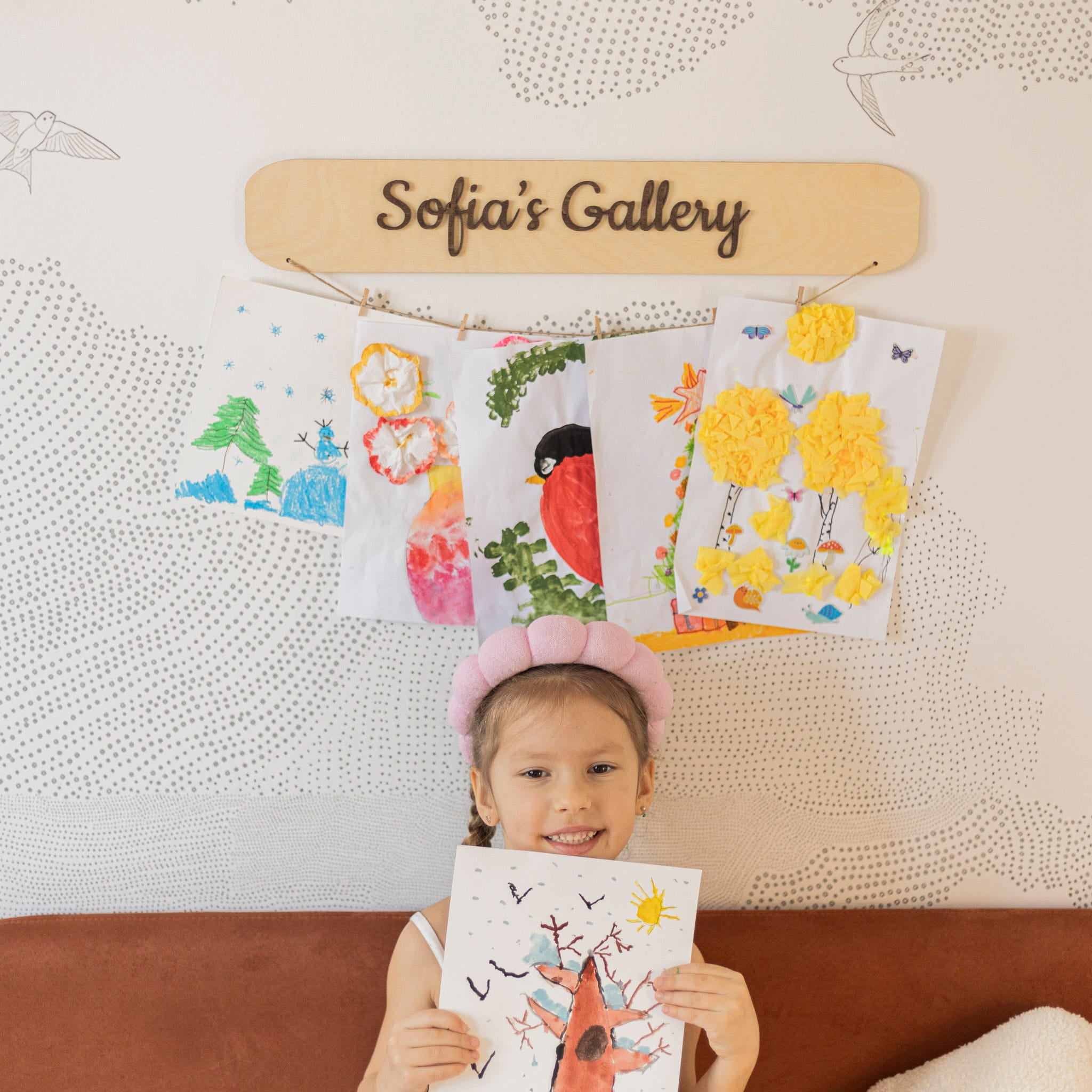 Child holding a drawing in front of a wall with children's artwork and a sign that reads 'Sofia's Gallery'.