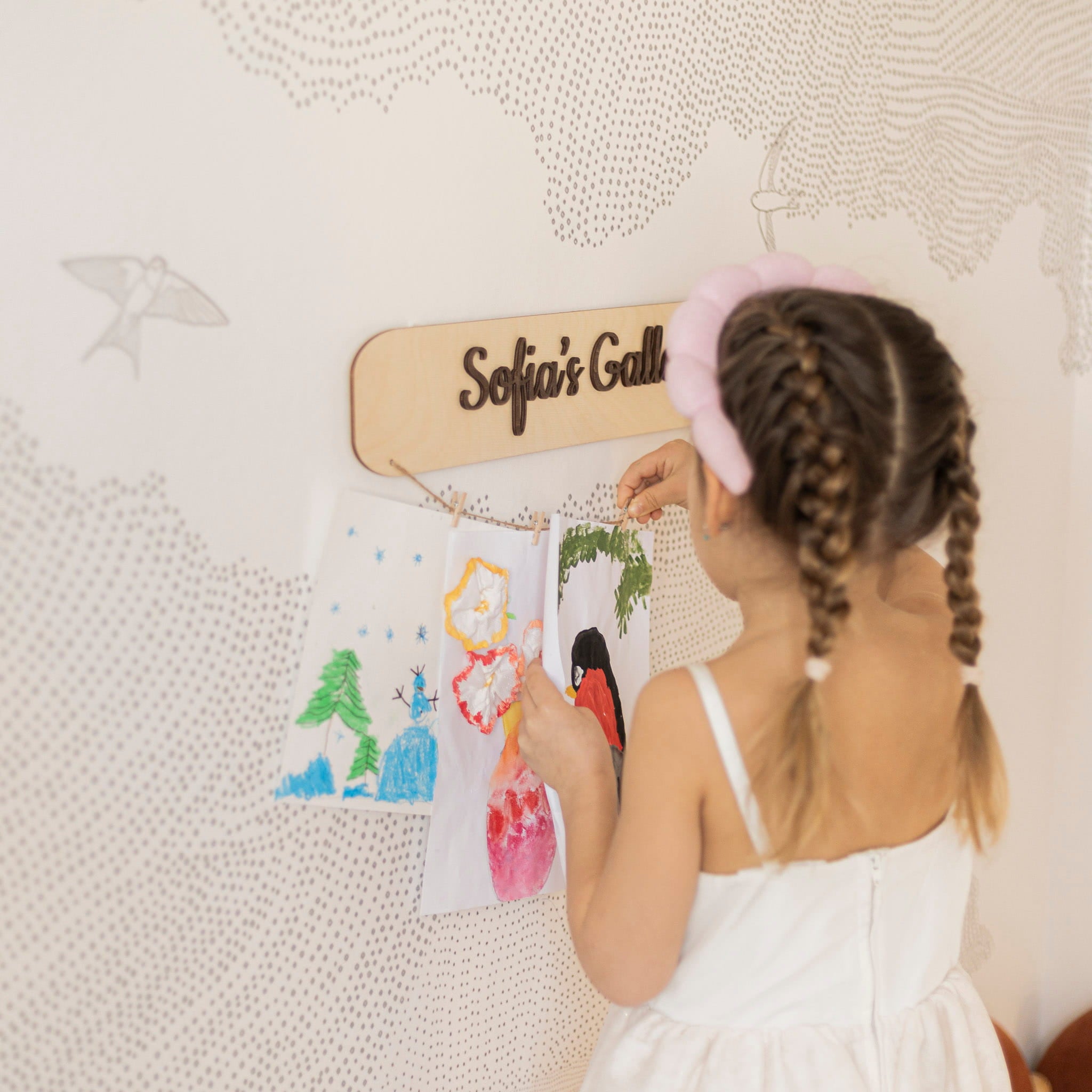 Child with braided hair and a pink hair accessory looking at drawings on a wall-mounted board.