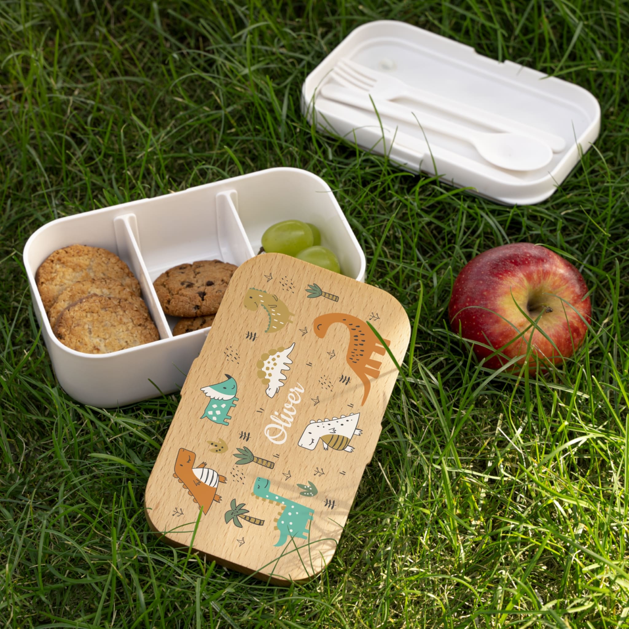 White bento box with compartments containing cookies and grapes, wooden lid with animal design, on grass.