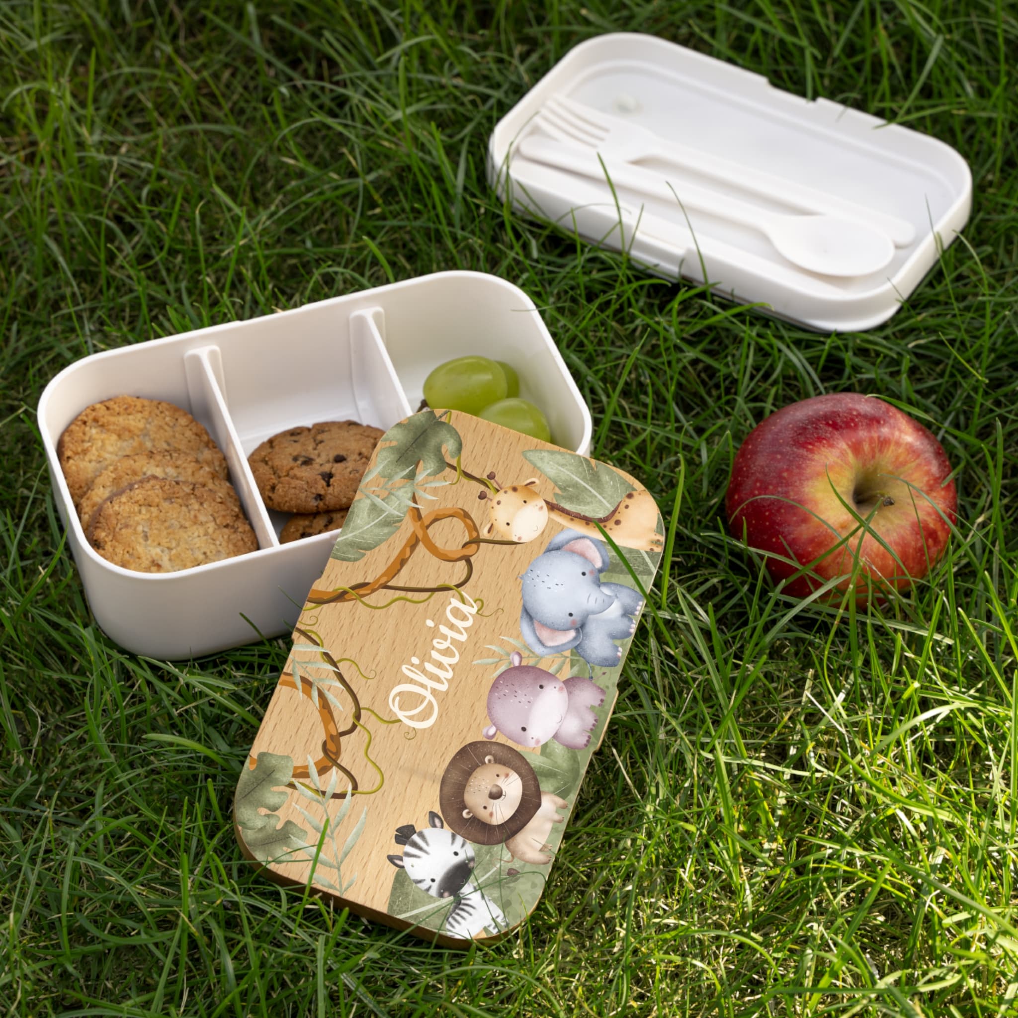 White bento box with compartments on grass, containing cookies and grapes, with a wooden nameplate featuring animal illustrations.