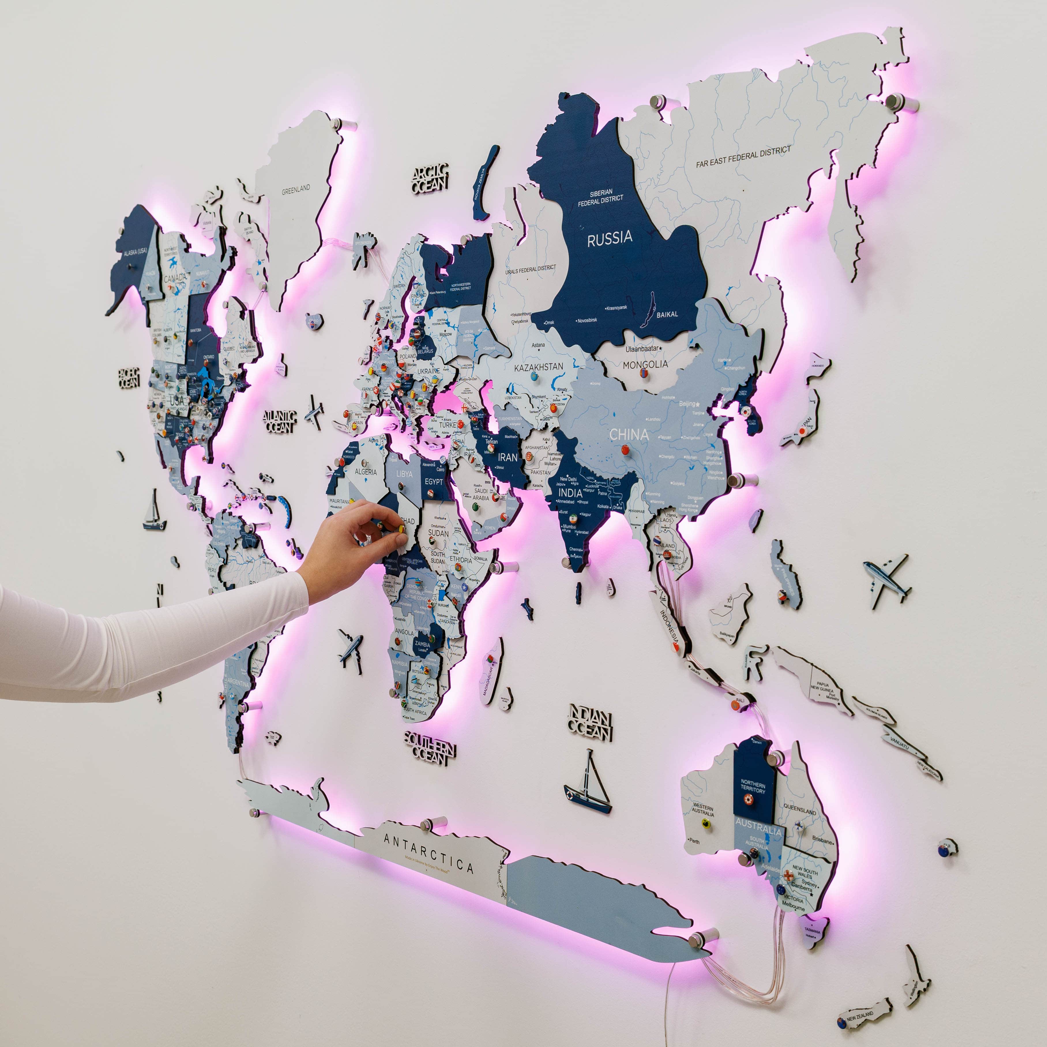 World map with LED lighting on a white wall, hand pointing at a location.
