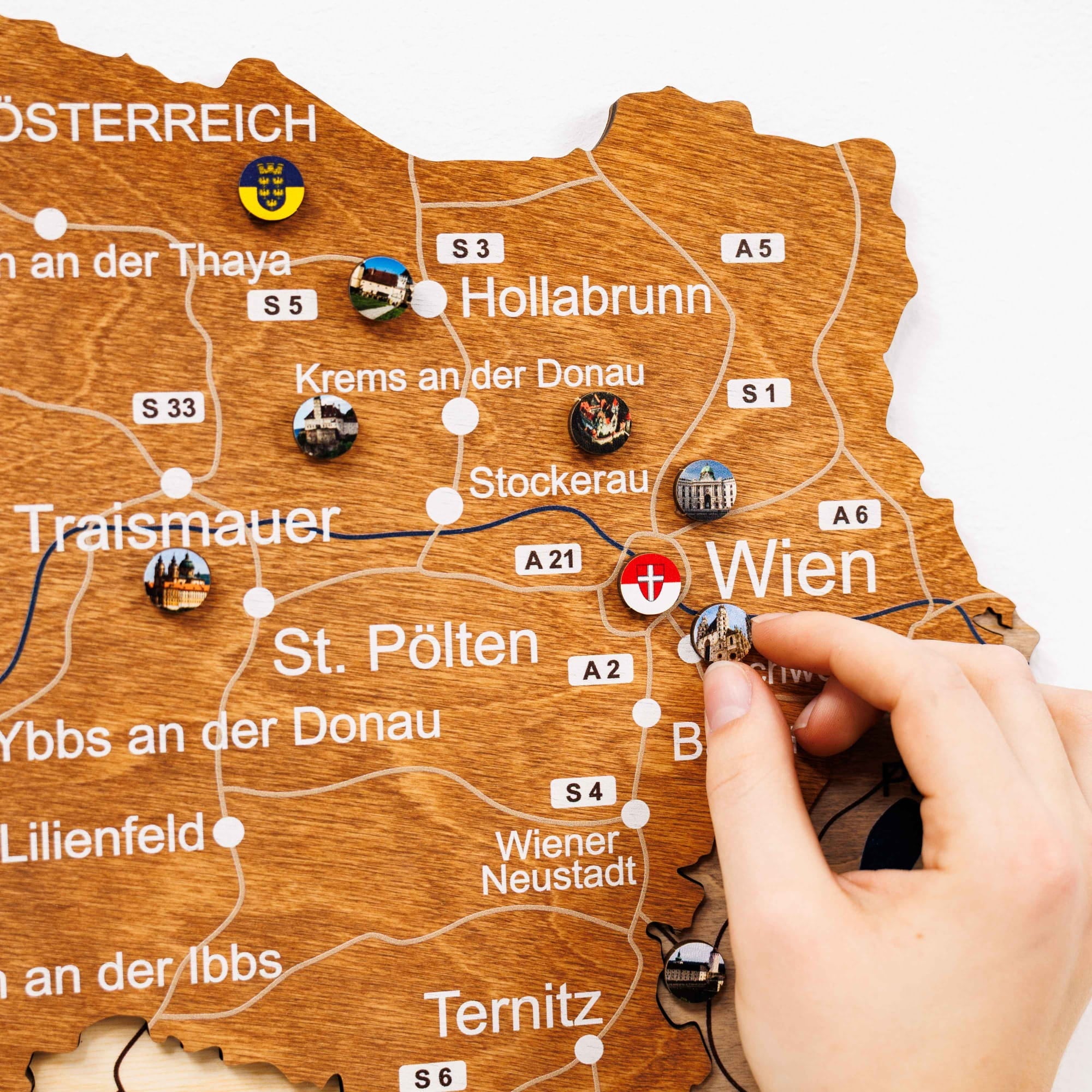 Wooden map of Austria with pins marking locations, hand placing a pin on 'Wien'.