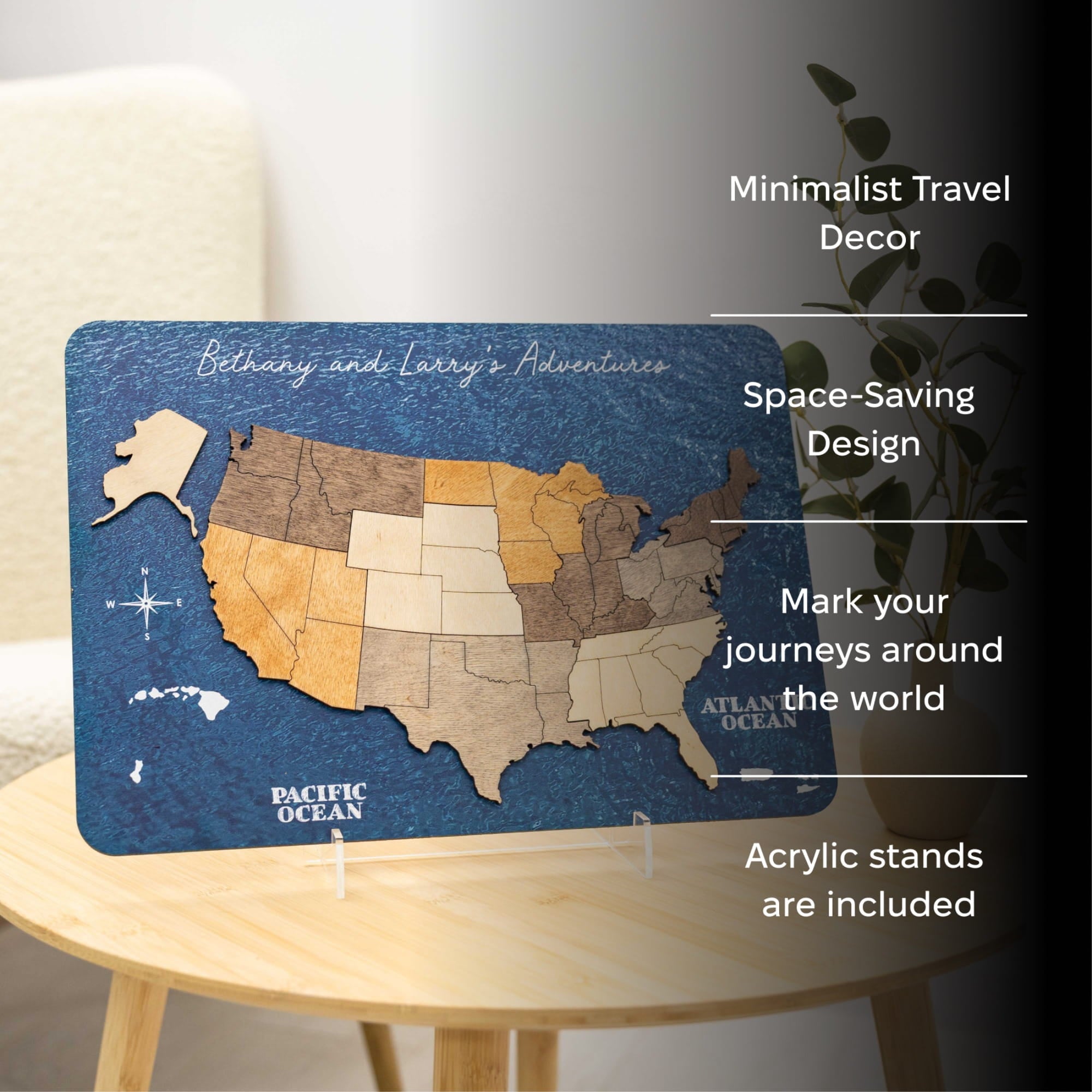 A rectangular acrylic travel wall decor plaque showing a stylized map of the United States with the title 'Bethany and Larry's Adventures' on a dark blue background. The plaque is displayed on a small round wooden table in a softly lit interior setting, with white text callouts on the right describing features like minimalist travel decor, space-saving design, mark your journeys around the world, and included acrylic stands.