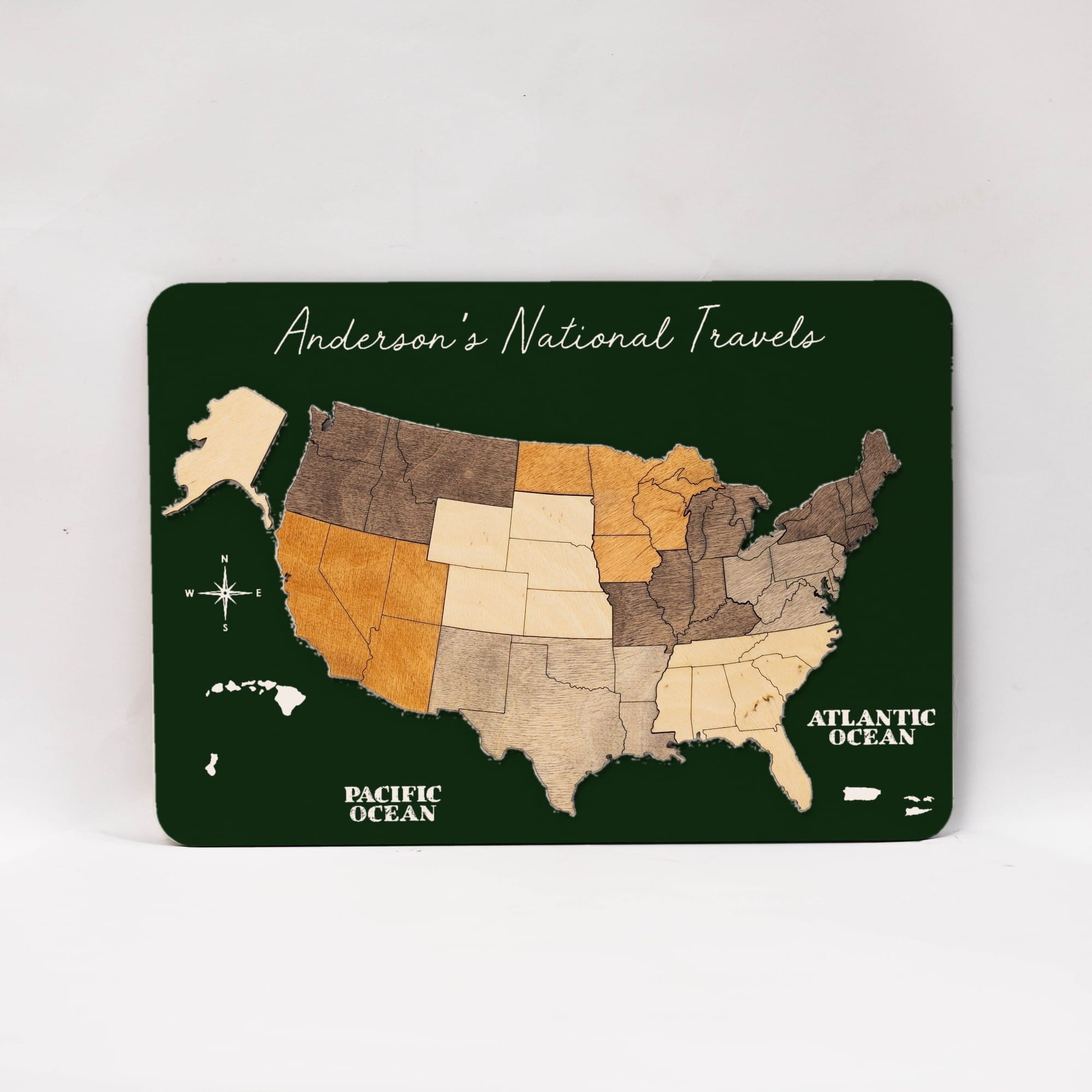 A rectangular dark green map plaque featuring a stylized United States with states in different wood-tone shades, labeled 'Anderson's National Travel' and 'Pacific Ocean' and 'Atlantic Ocean,' photographed on a light neutral background.