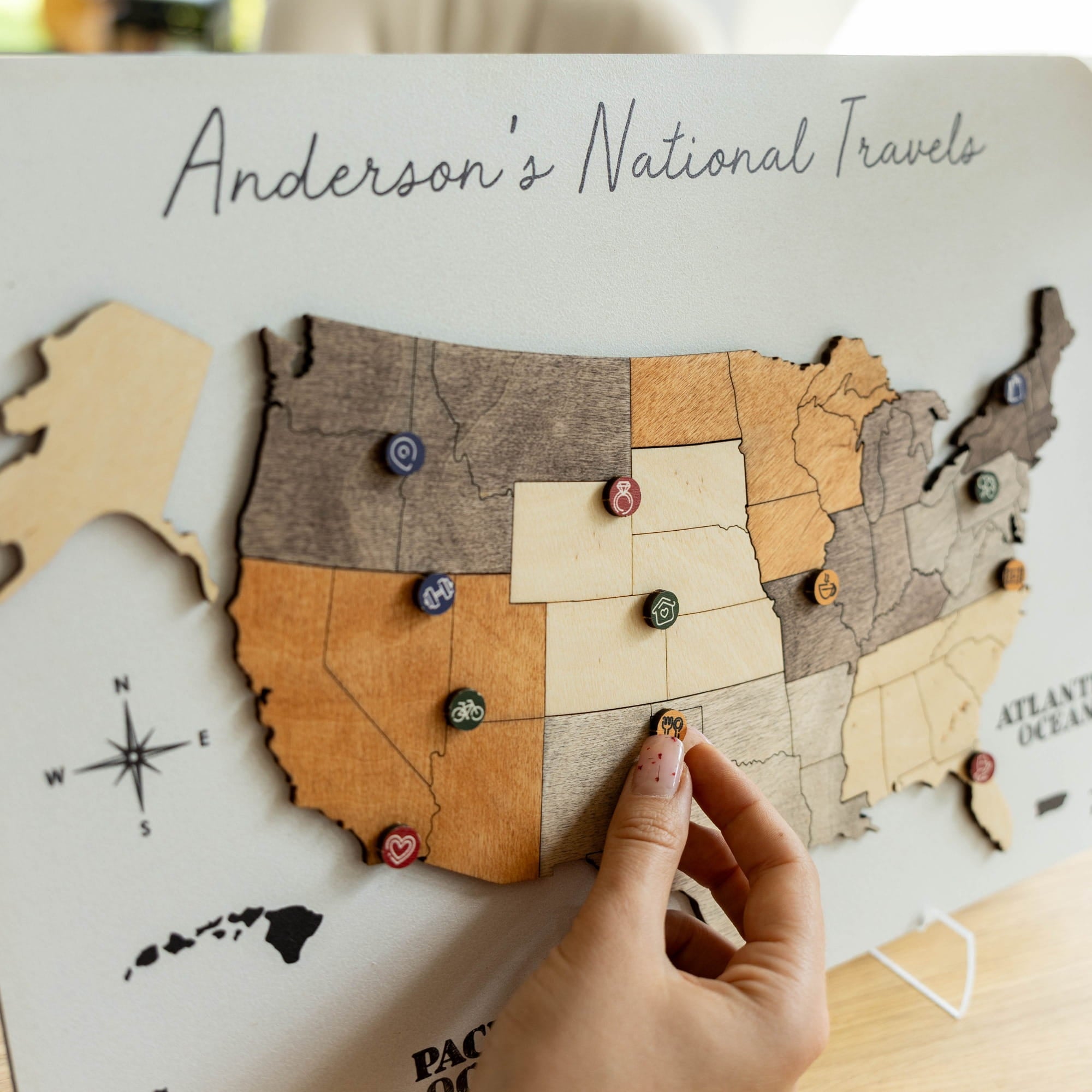 A wooden US map travel board labeled 'Anderson's National Travels,' with a hand placing a small decorative marker on a state section. The board shows multiple states in natural wood tones with small colorful travel pins.