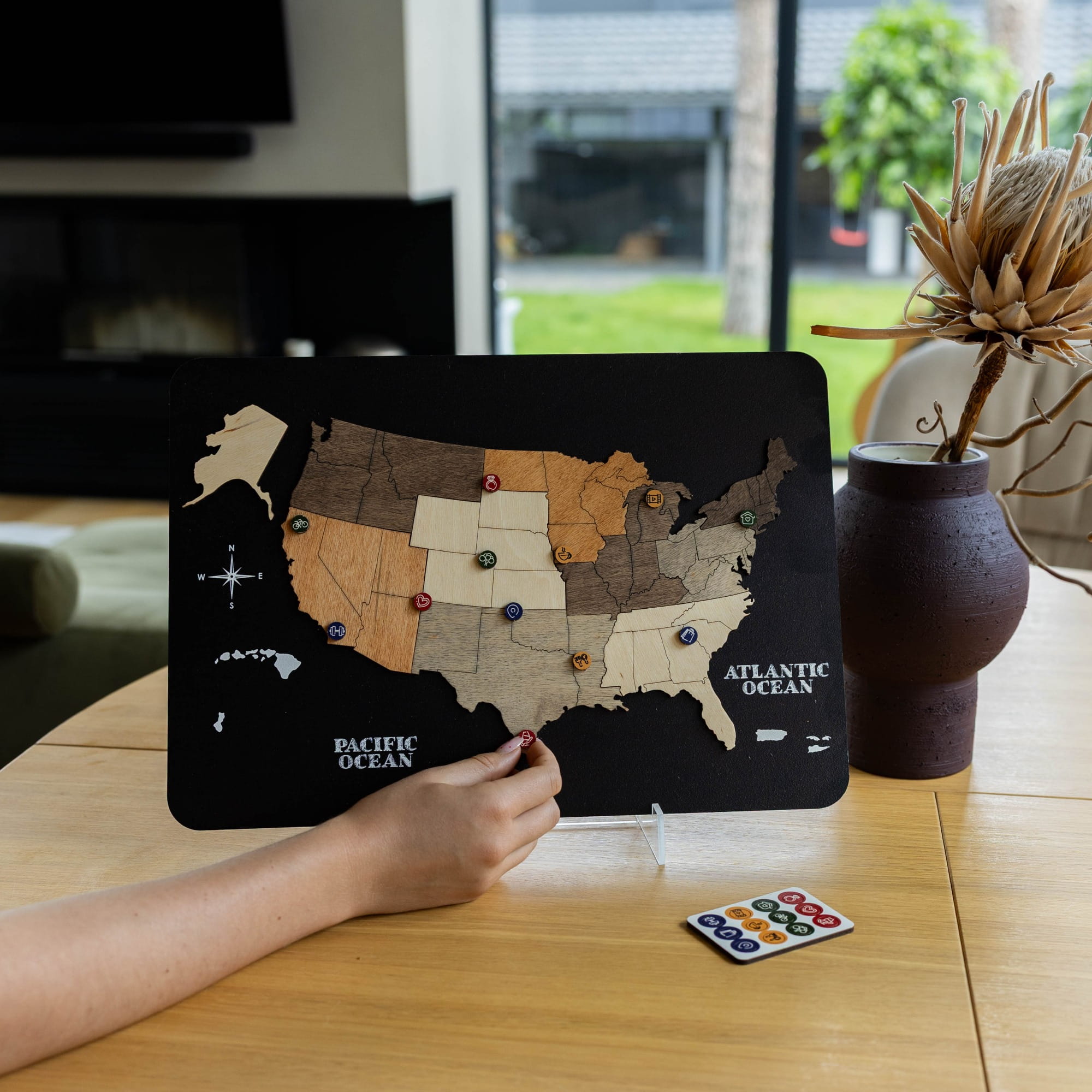 A person's hand is shown interacting with a black magnetic US map board game on a wooden table, with a small remote control nearby and a dark brown ceramic vase with dried stems in the background.