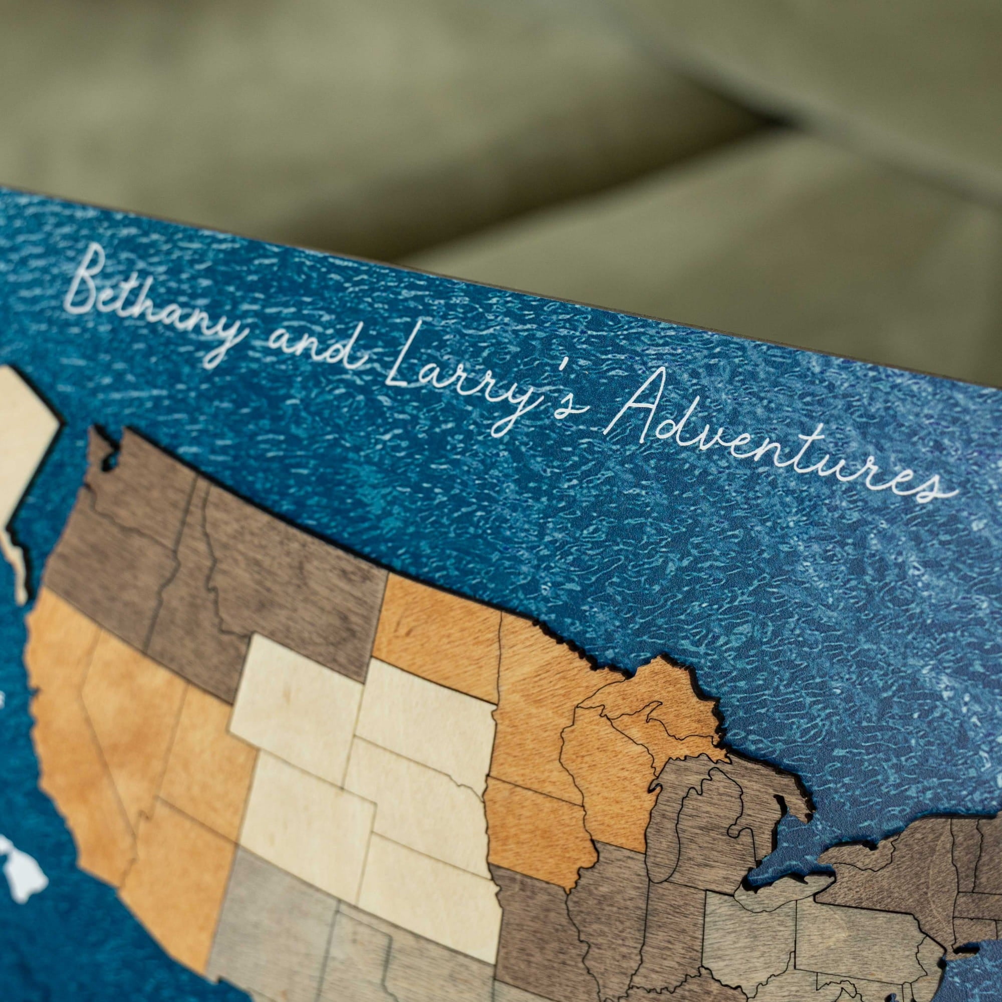 Close-up of a wooden map plaque featuring the Great Lakes region, with engraved states in different wood tones on a dark blue textured background. White script text reads 'Bethany and Larry's Adventures'.