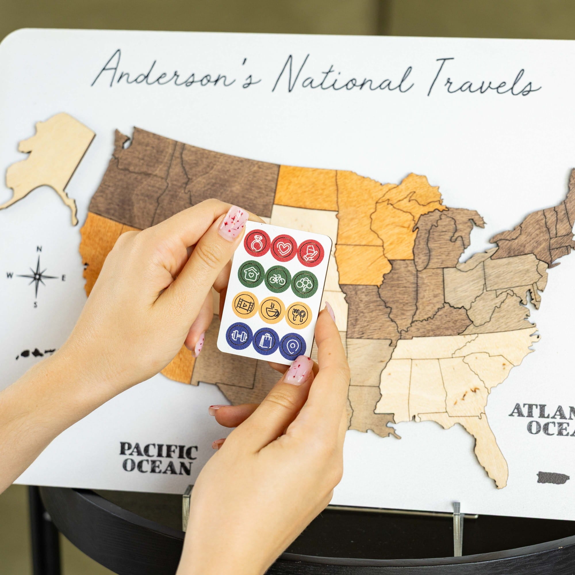 A person's hands holding a small white card with colorful round travel icons in front of a wooden US map labeled 'Anderson's National Travels.'.