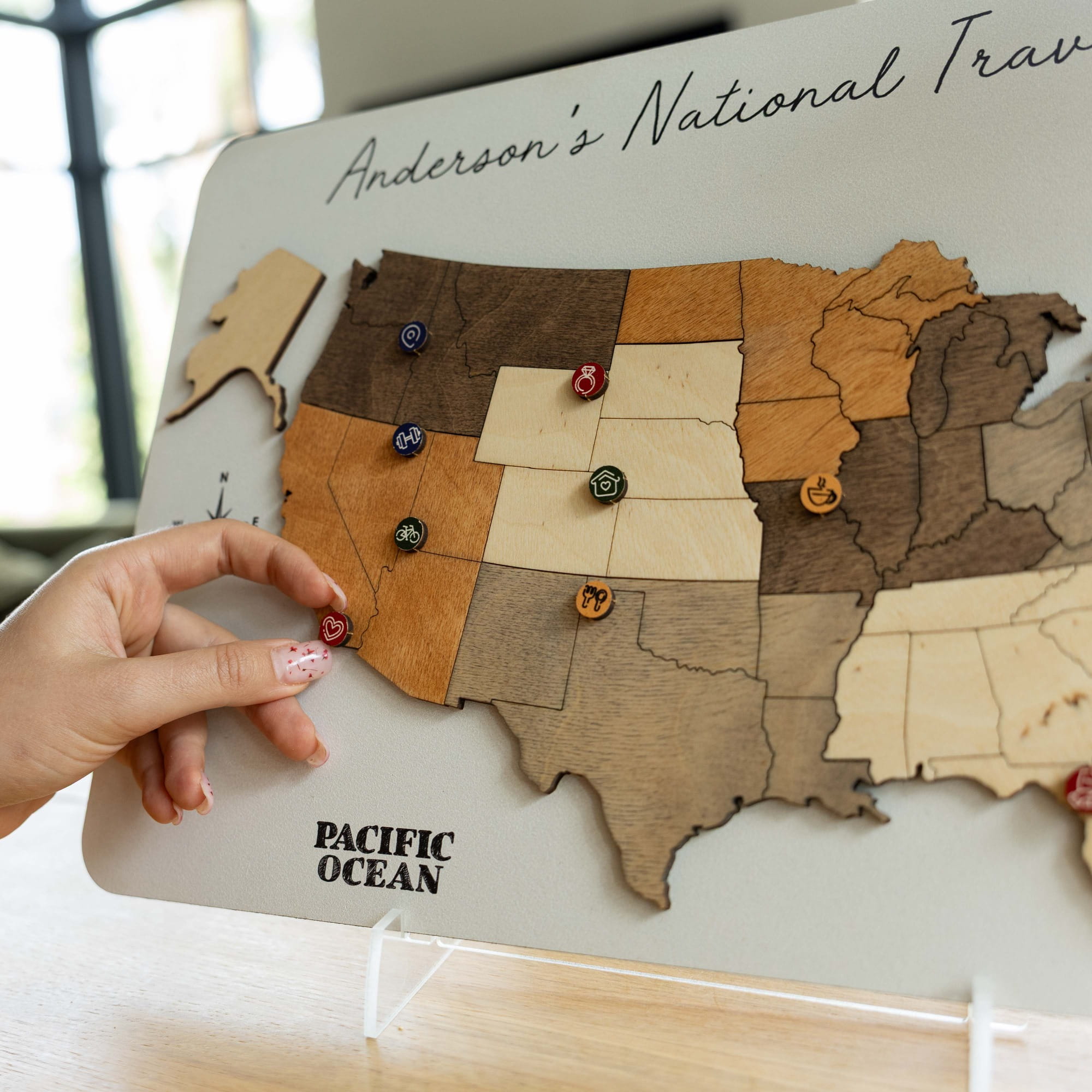 A close-up of a wooden US state travel map board with a hand placing a small travel token on California. The board has a light gray base, natural and stained wood states, and the text 'Anderson's National Travel' at the top and 'PACIFIC OCEAN' at the bottom.