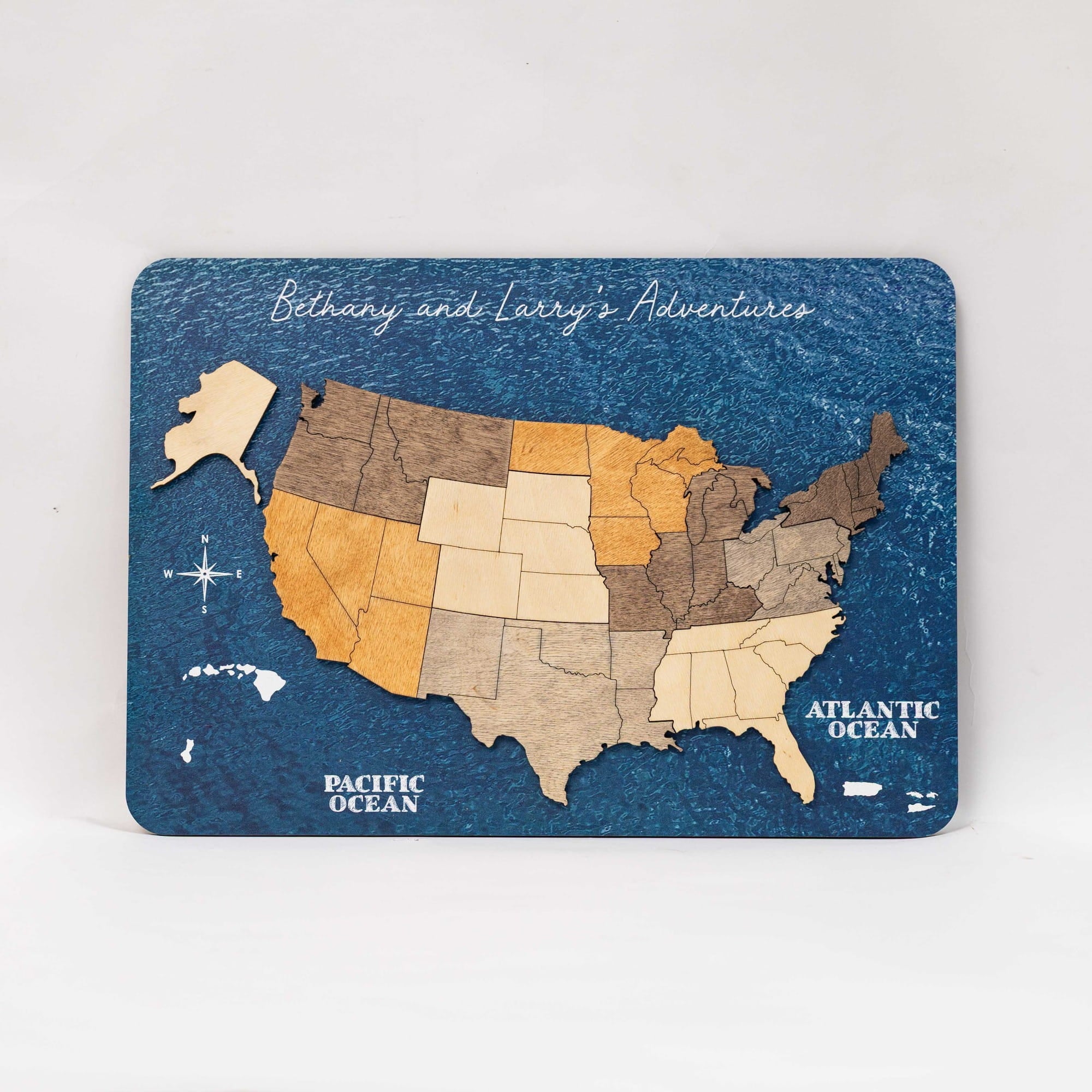 A rectangular wooden map plaque of the United States titled 'Bethany and Larry's Adventures' with states cut from natural wood in various tones, set against a textured dark blue background representing the oceans.