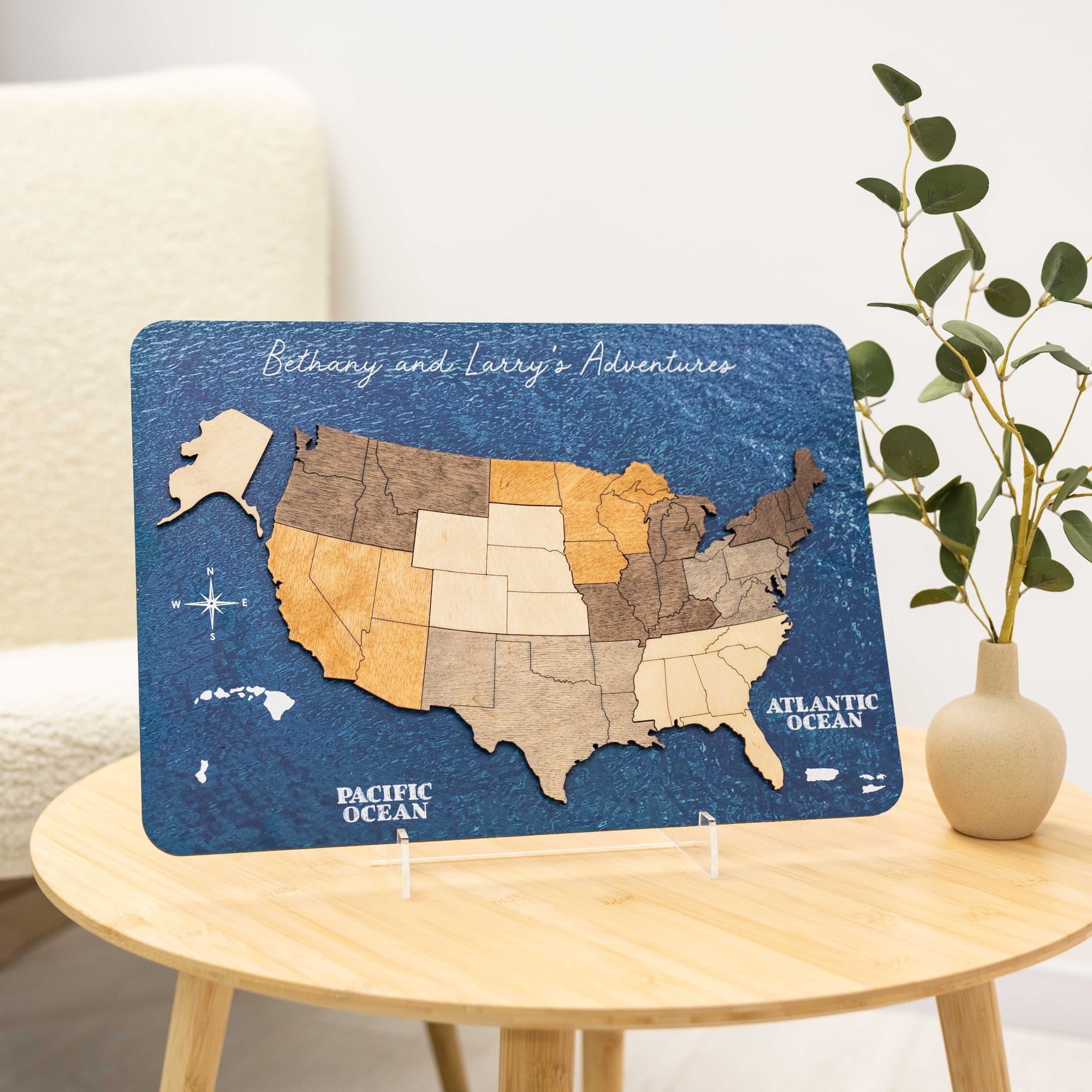 A rectangular wooden US map plaque with a textured blue background and state sections in different wood tones, displayed on a small round wooden table in a styled interior setting with a vase of greenery.