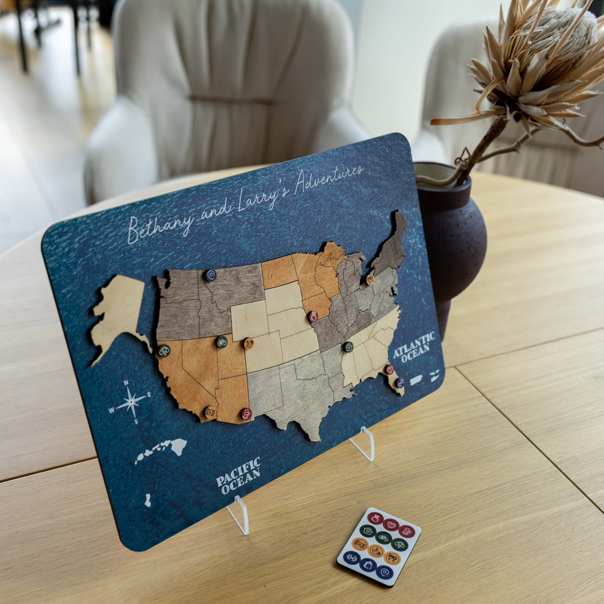 A wooden US states travel map plaque with a dark blue ocean background and state cutouts, displayed on a tabletop with small colorful pins and a remote control. The scene includes a chair and vase in a real indoor environment, so this is Lifestyle rather than Product/Packaging.