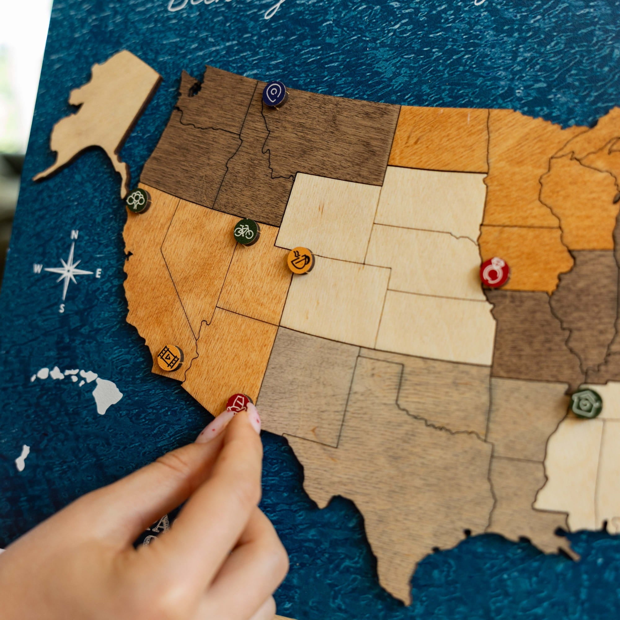A close-up of a wooden United States map with states carved in different wood tones, set against a dark blue textured background. A person's hand is visible at the bottom left, touching a small red pin on Texas, while several decorative wooden pins are placed across the states.