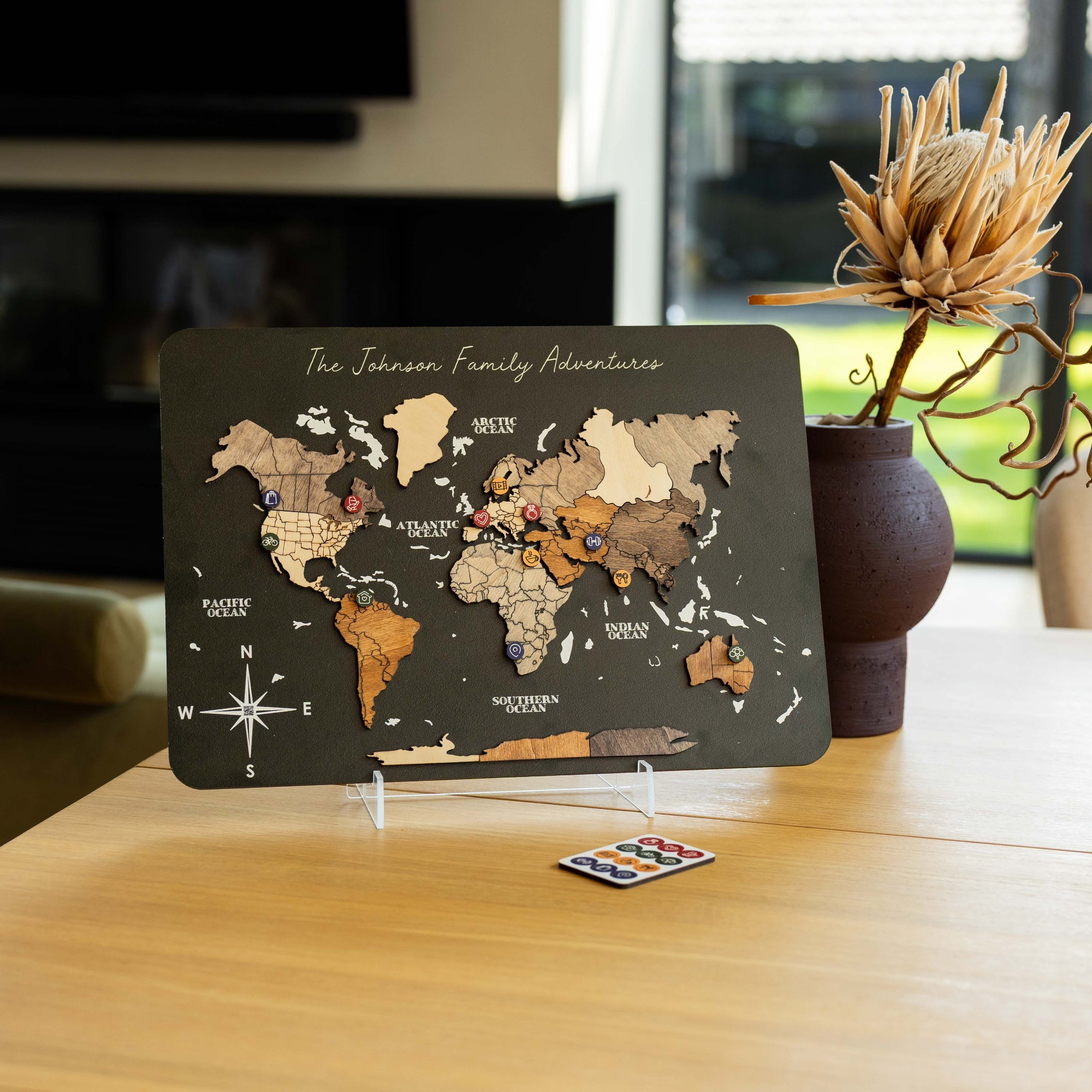 A wooden world map travel plaque with raised continents and the text 'The Johnson Family Adventures' displayed on a clear stand on a wooden table, with a small compass and a dried decorative plant in the background.