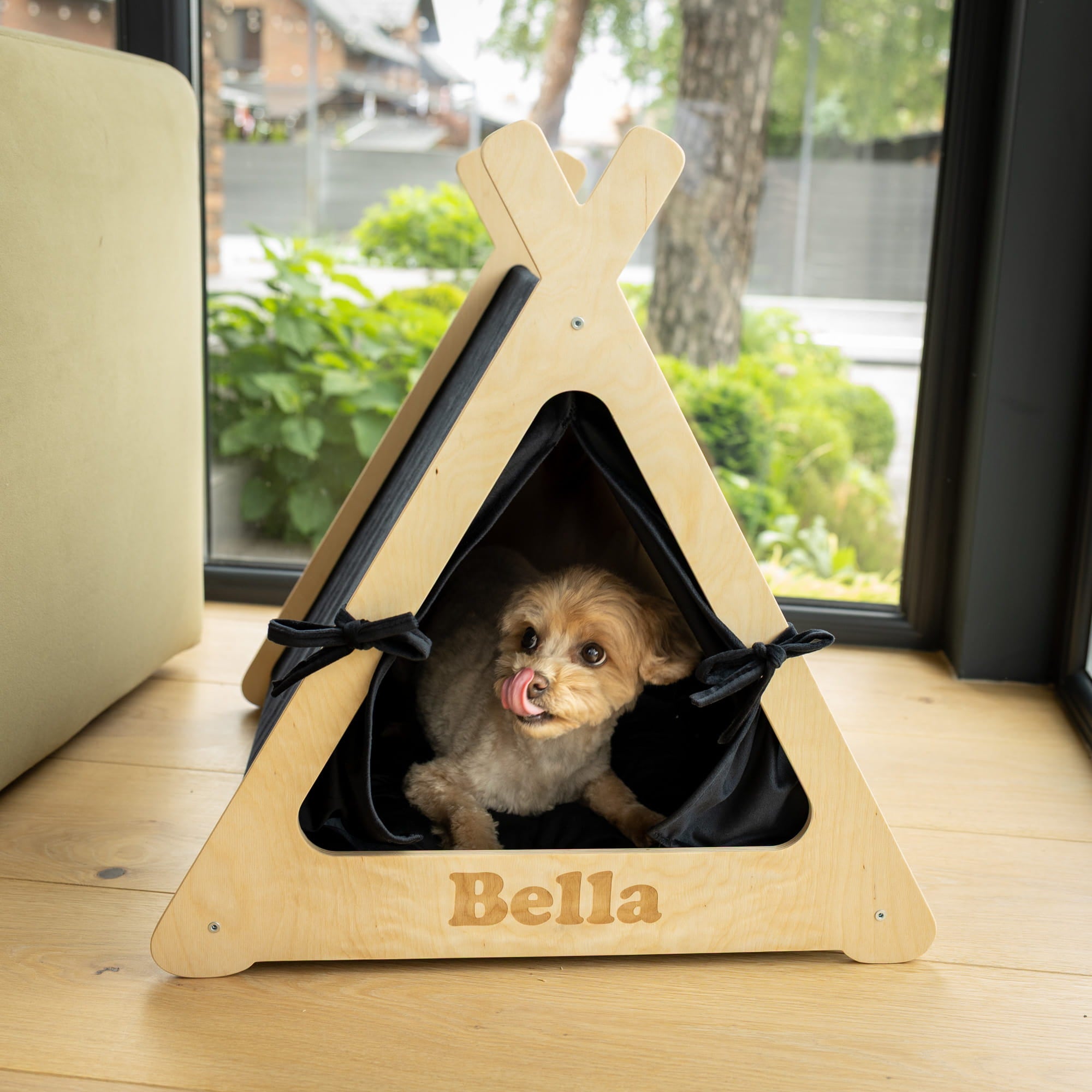 Dog peeking out from a wooden pet teepee named 'Bella' on a wooden floor.