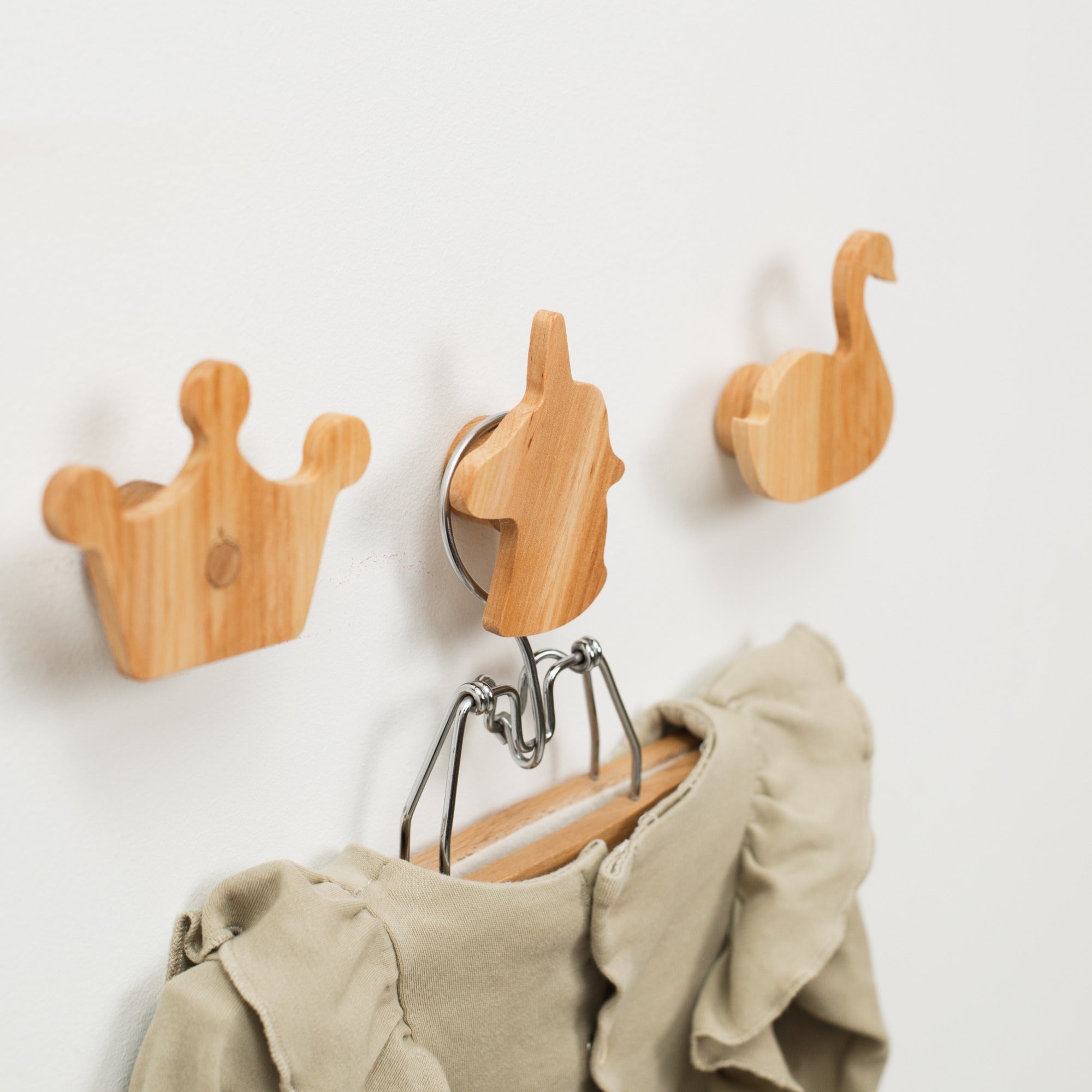 Wooden wall hooks shaped like a crown, horse, and swan with a beige garment hanging from one of them on a light gray background.