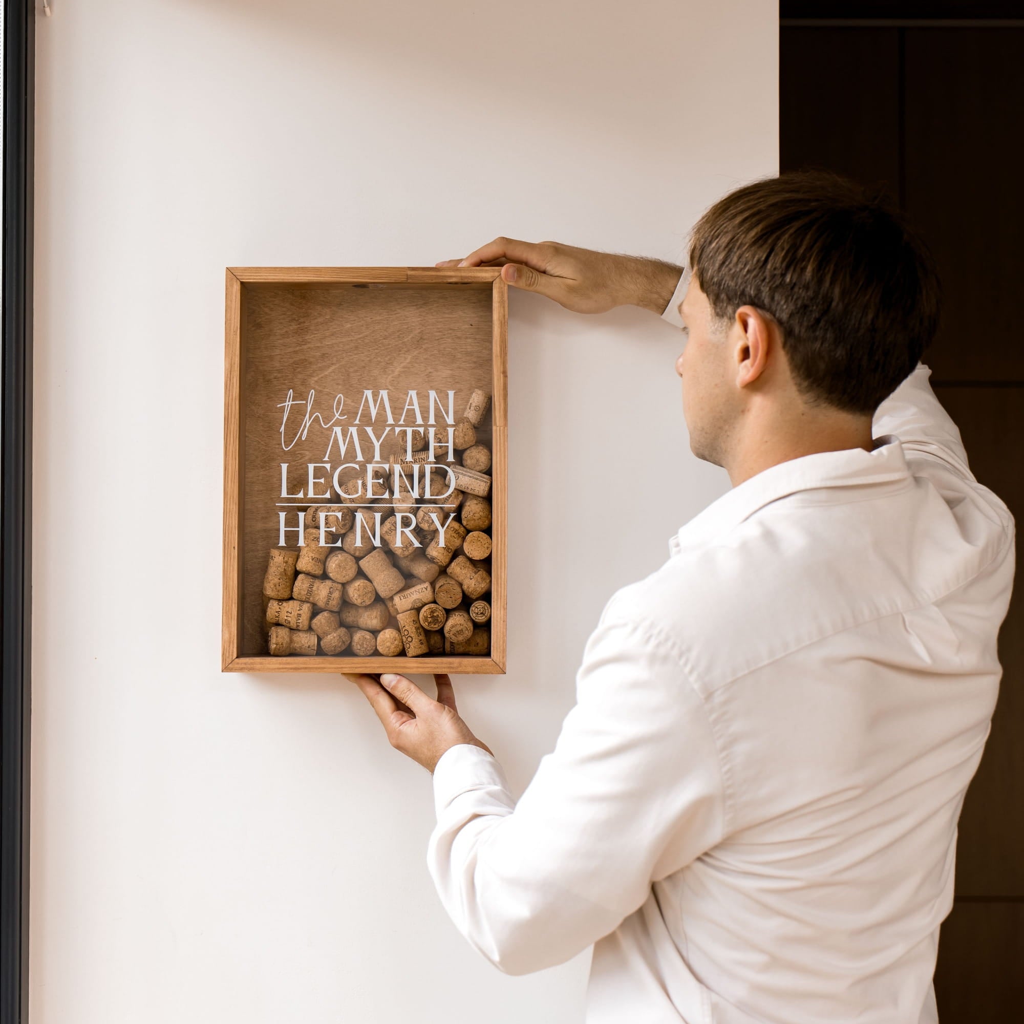 A man in a white shirt holds a wooden framed wall decor piece filled with wine corks and featuring the text 'the MAN MY TH LEGEND HENRY' on a plain off-white wall.