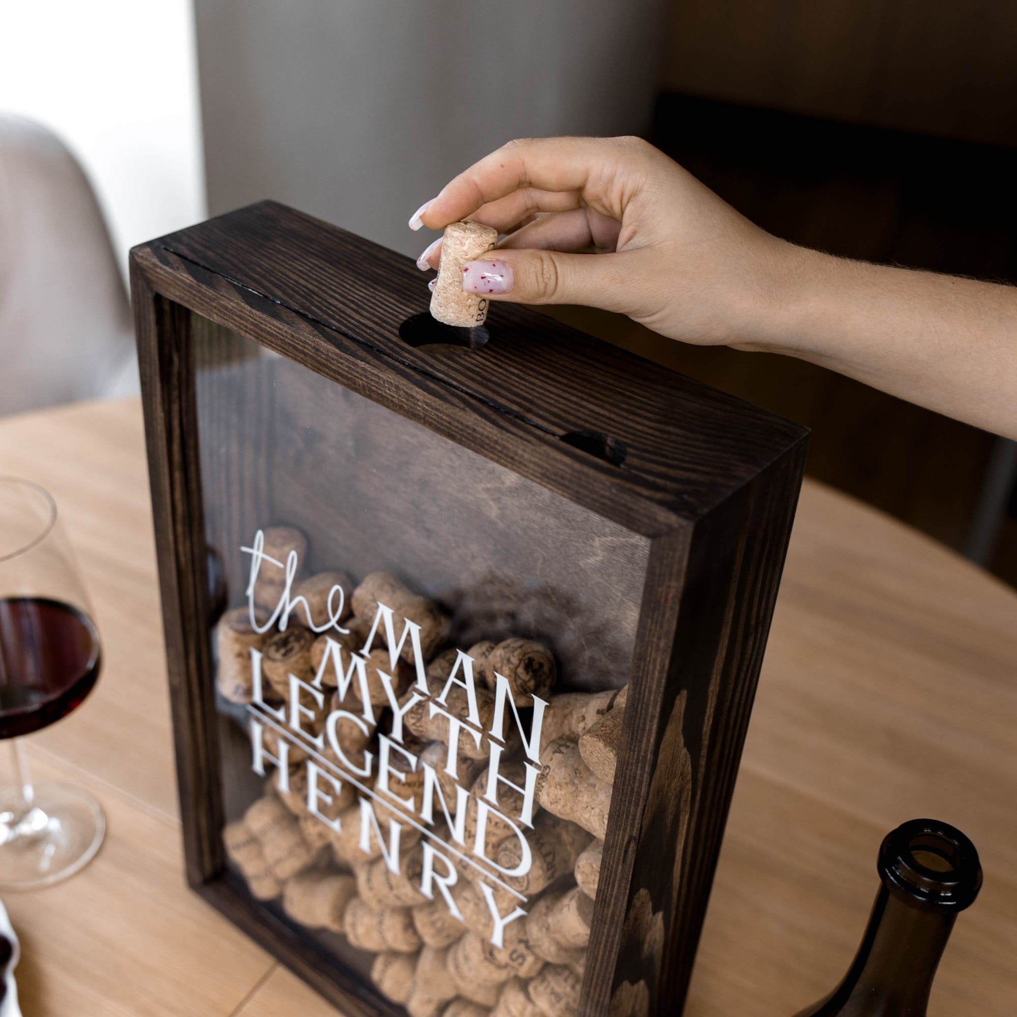 A hand inserts a wine cork into a dark wooden framed box