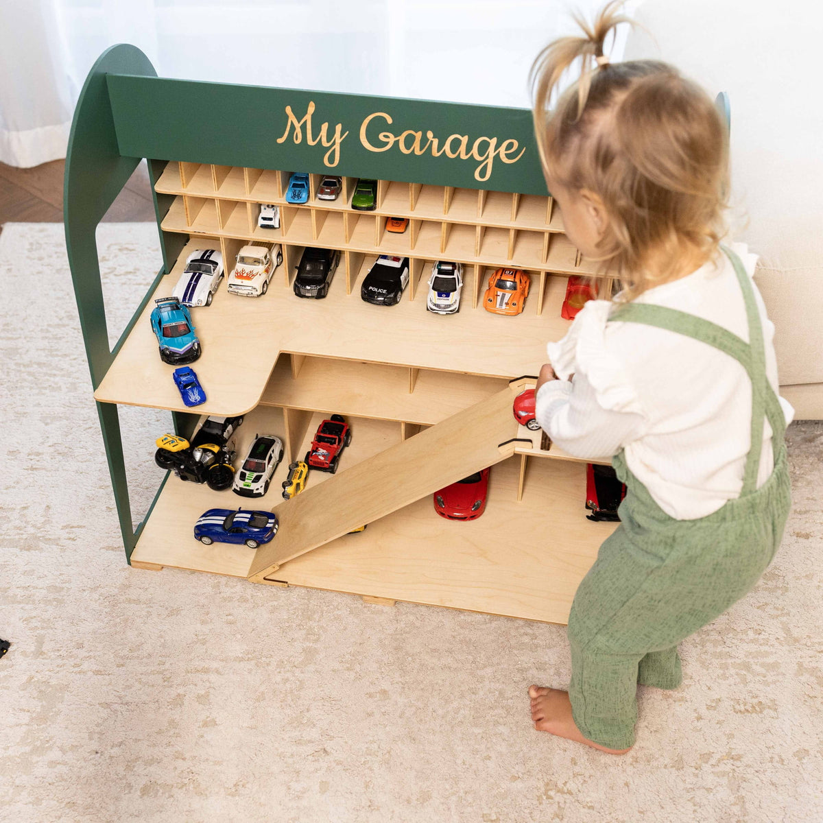 The garage has a dark green exterior, a light wood interior, a ramp, and multiple toy cars.