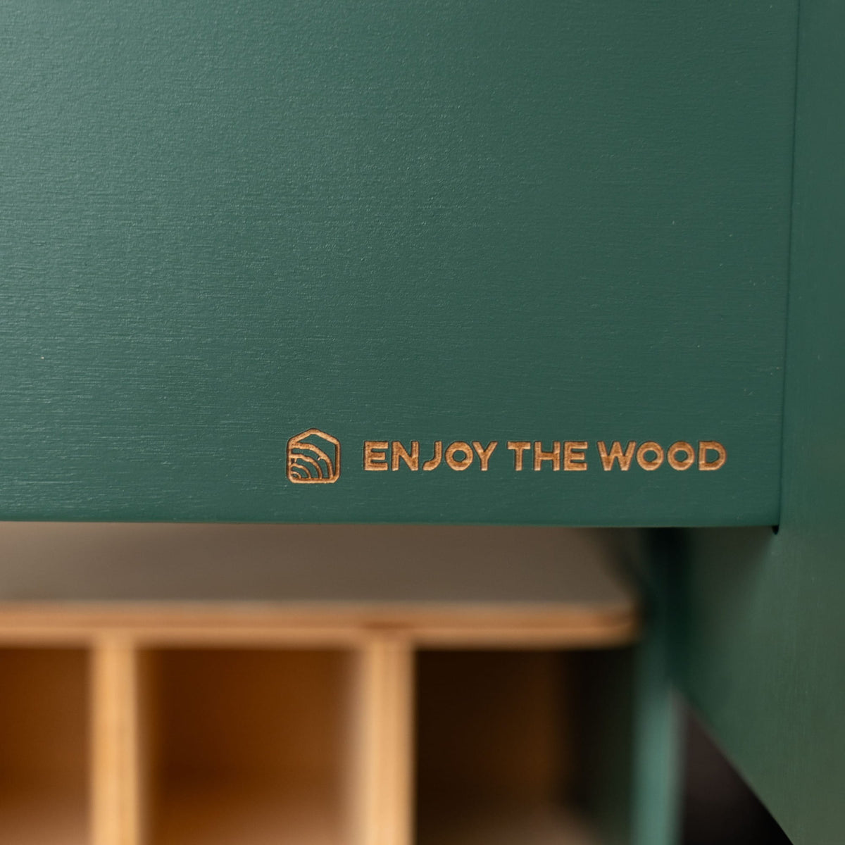 Close-up of a dark teal-green wooden cabinet or shelf with a gold foil logo that reads 'ENJOY THE WOOD' and a small geometric icon, with a blurred light wood shelf unit below.