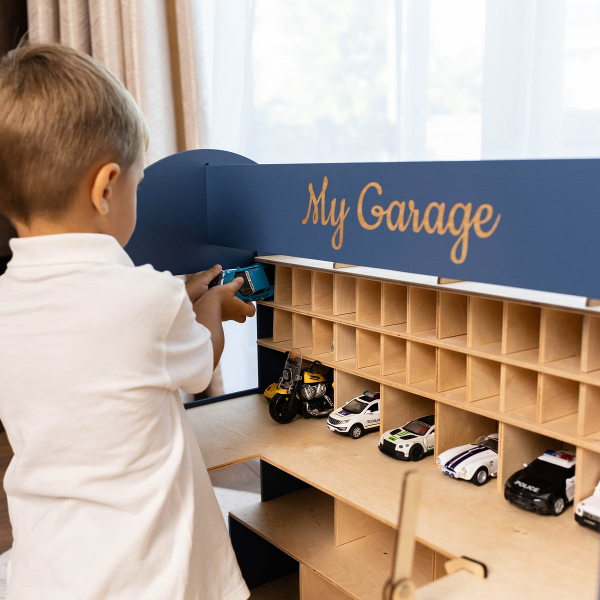 The garage has multiple light wood compartments holding small toy cars and trucks, set in a bright room near a window.