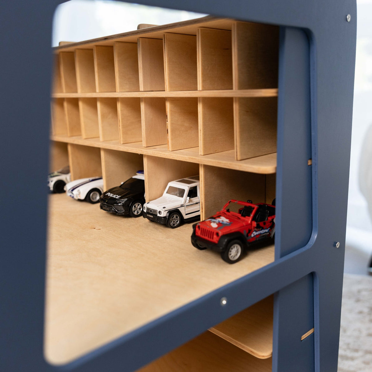 A close-up view of a wooden toy car garage shelf with cubby compartments, some filled with small toy cars including a red Jeep-style vehicle, photographed through a blue frame.