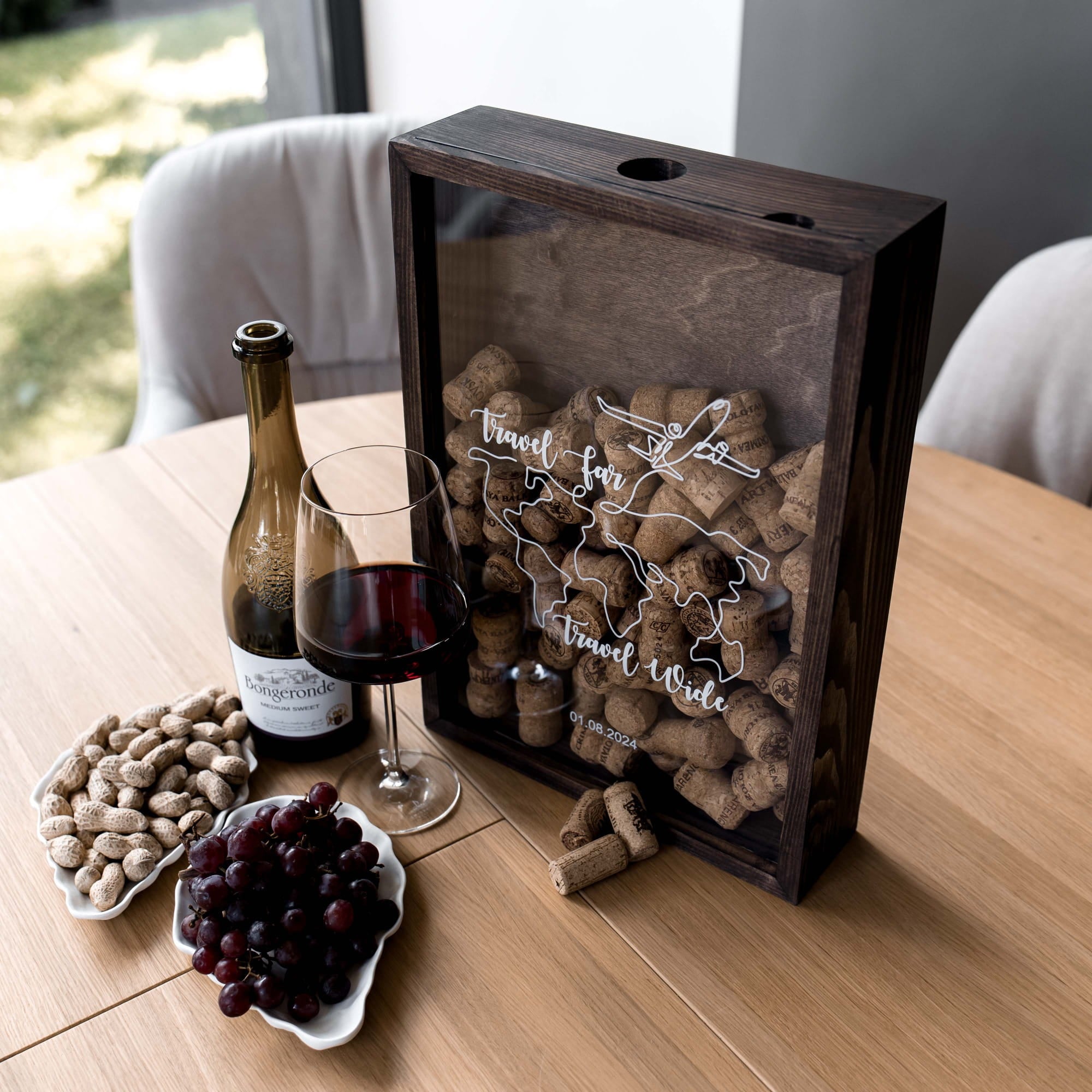 A wooden shadow box wine gift set on a light wood table, featuring a dark wood frame, a clear front panel with a white map outline and 'Travel Far Wine' script, filled with cork stoppers. 