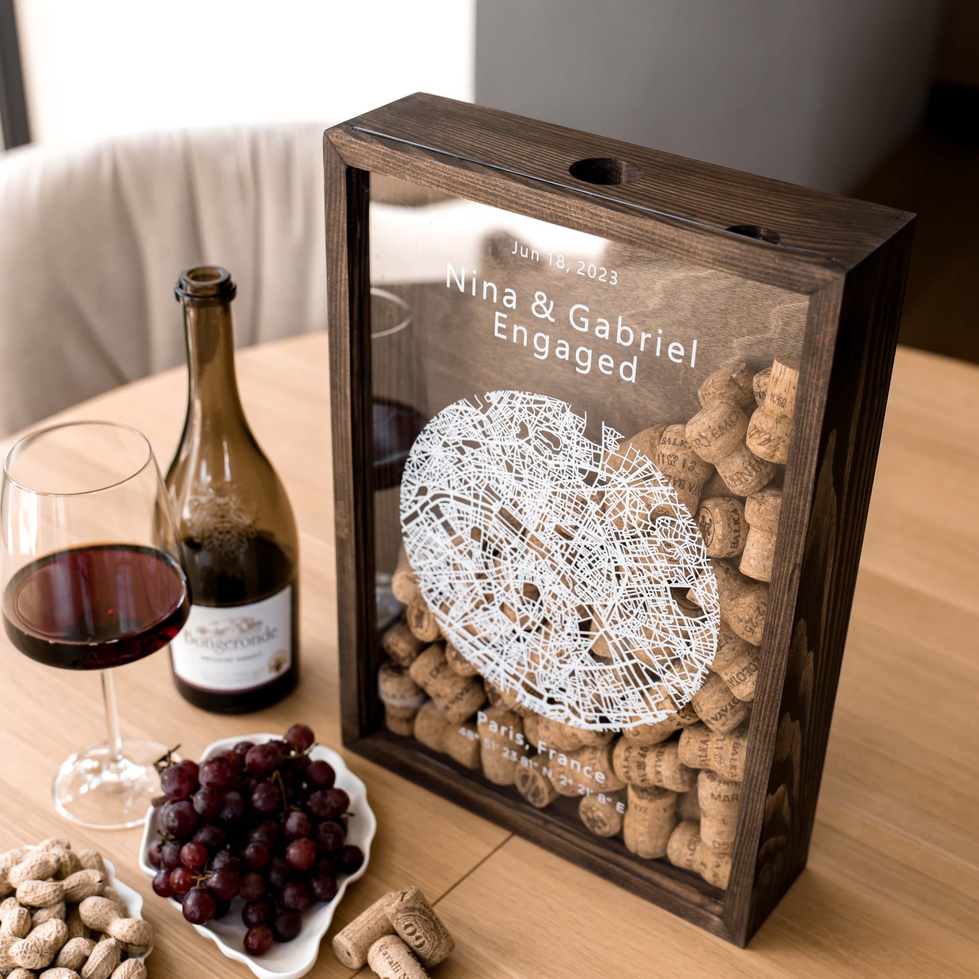 A personalized wooden wine gift box with a glass front panel sits on a wooden table.