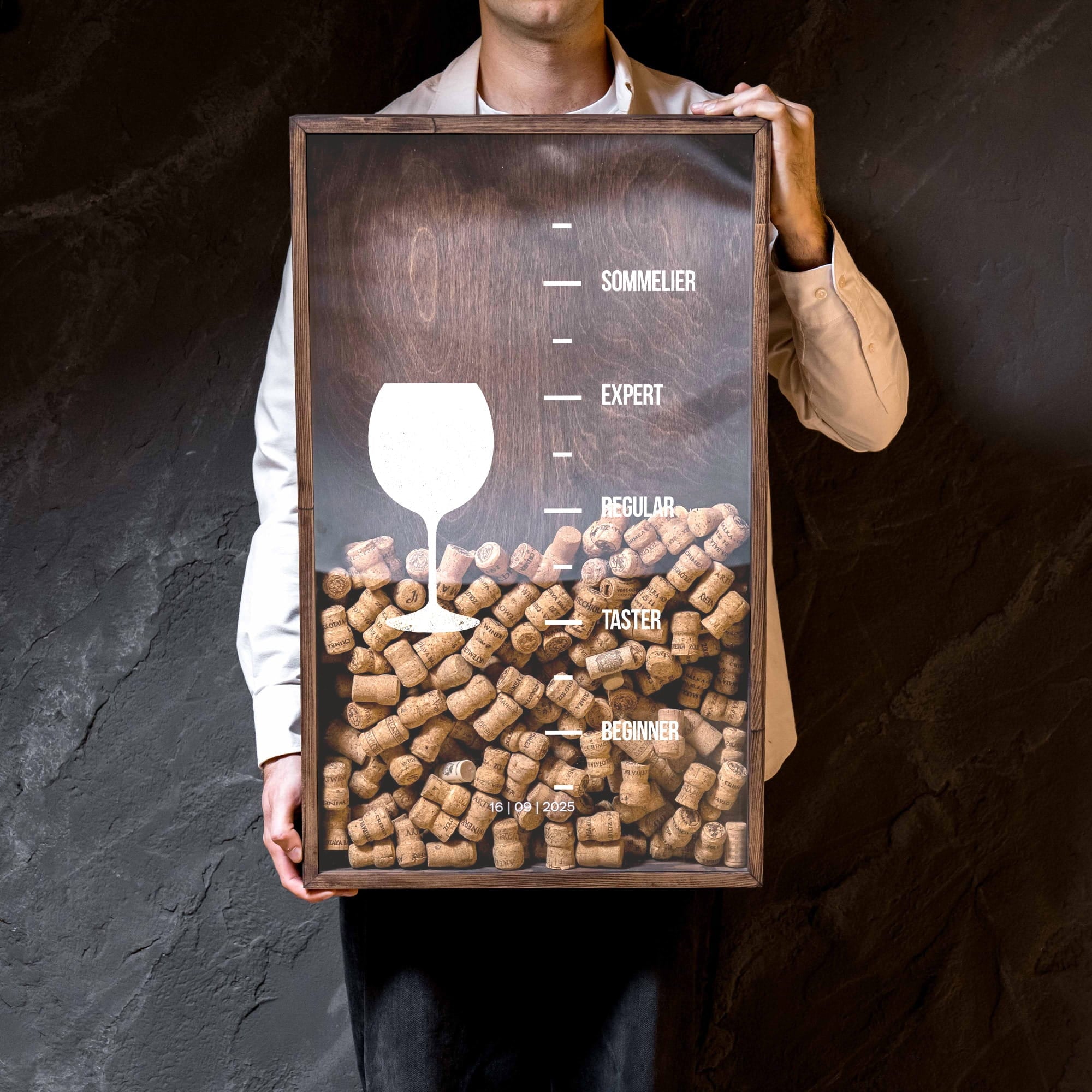 A person in a white shirt and tan jacket holds a framed art print featuring a wine glass silhouette layered over stacked wine corks,