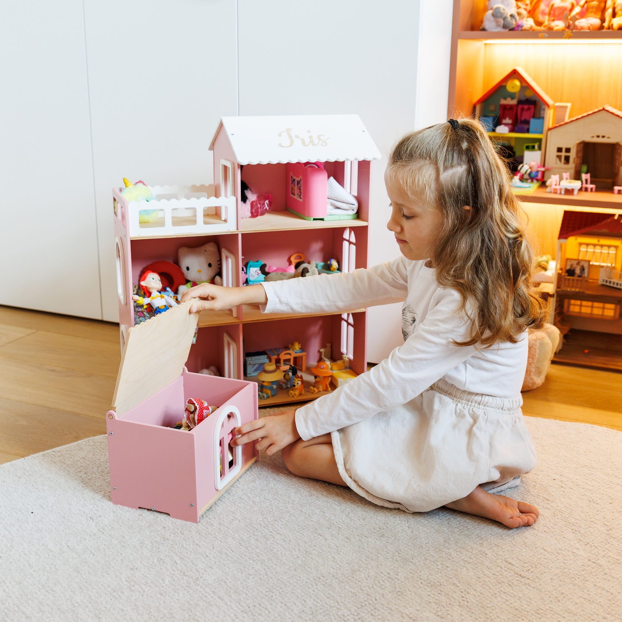 A young girl with long light brown hair sits on a light beige rug, playing with a pink wooden dollhouse and storage box in a brightly lit room.