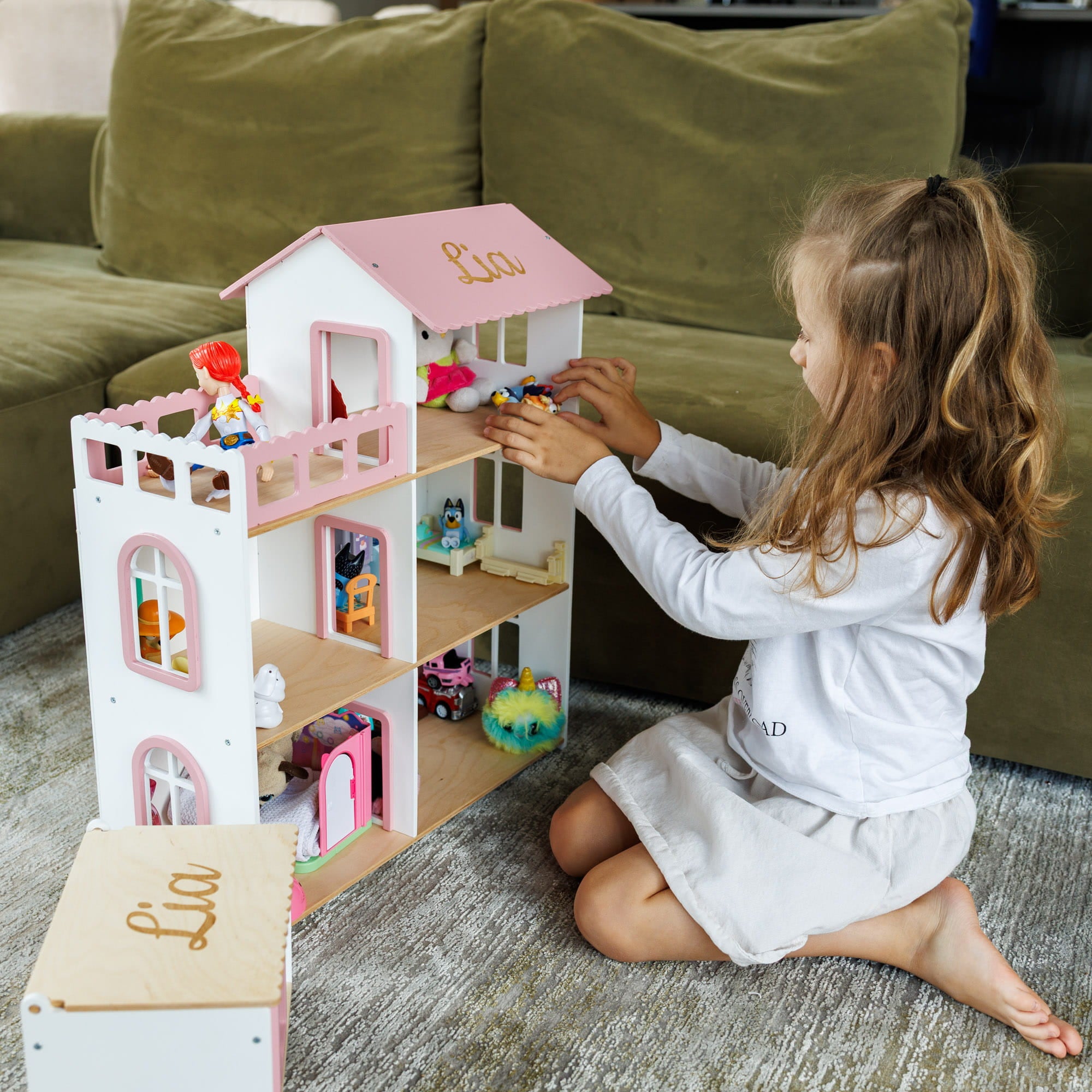 A young girl with long light brown hair sits on a gray carpeted floor, playing with a three-story wooden dollhouse.