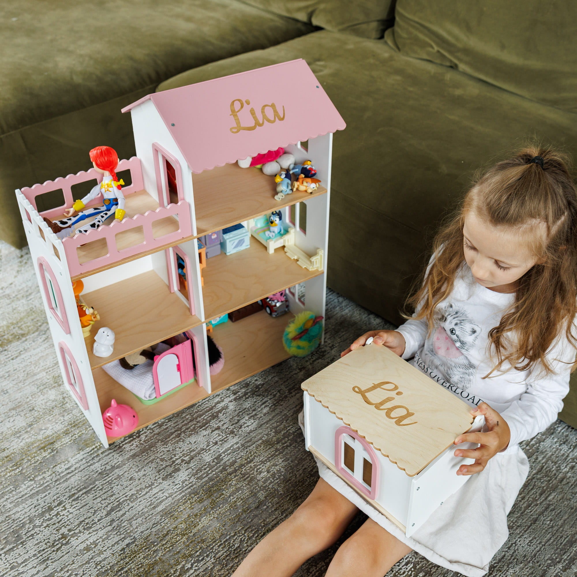 A young girl sits on a gray rug next to a large, personalized two-story wooden dollhouse with pink accents and the name 'Lia' engraved on the roof. 