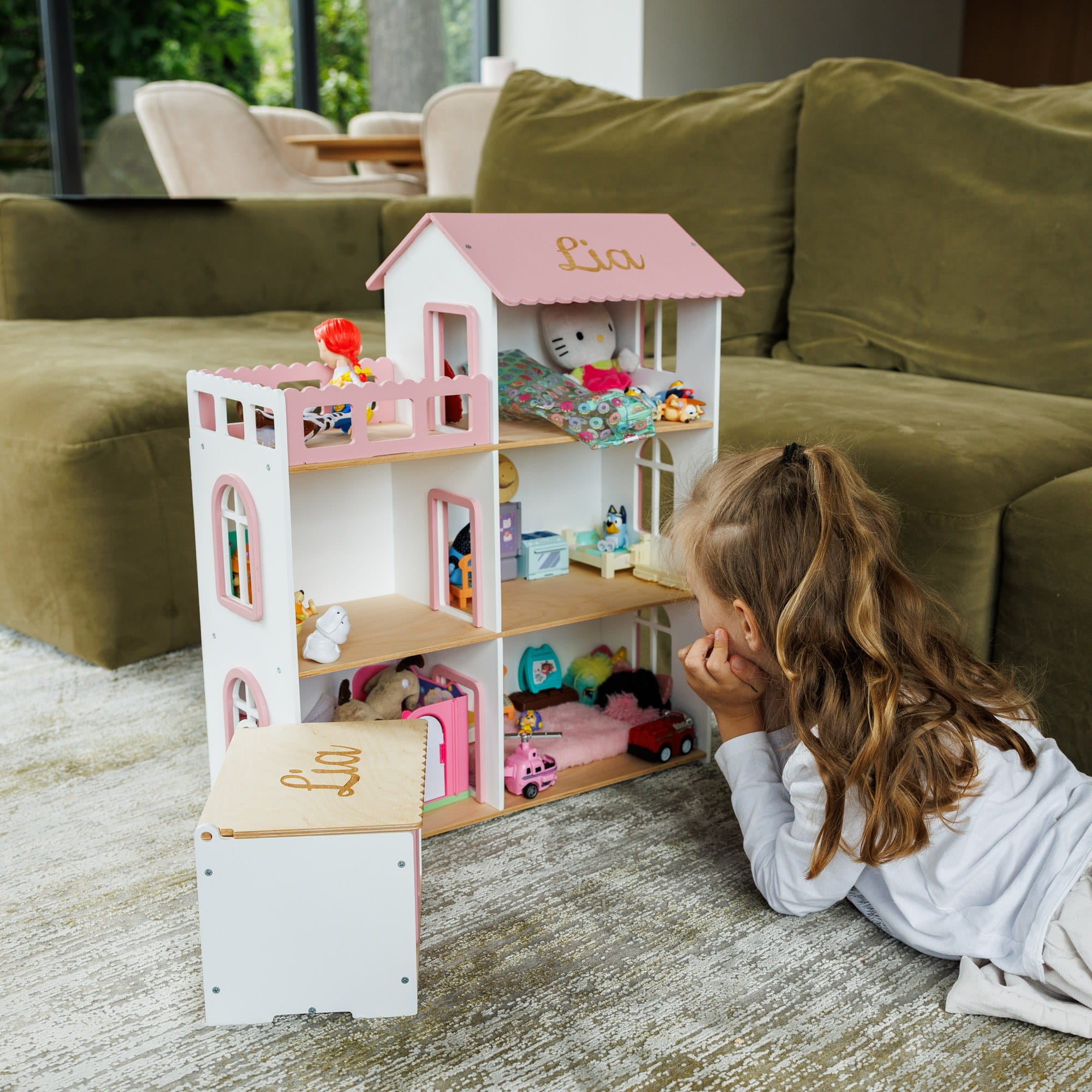 The dollhouse is white with pink trim, featuring a pink roof with the name 'Lia' in gold script, and is filled with various toy accessories.