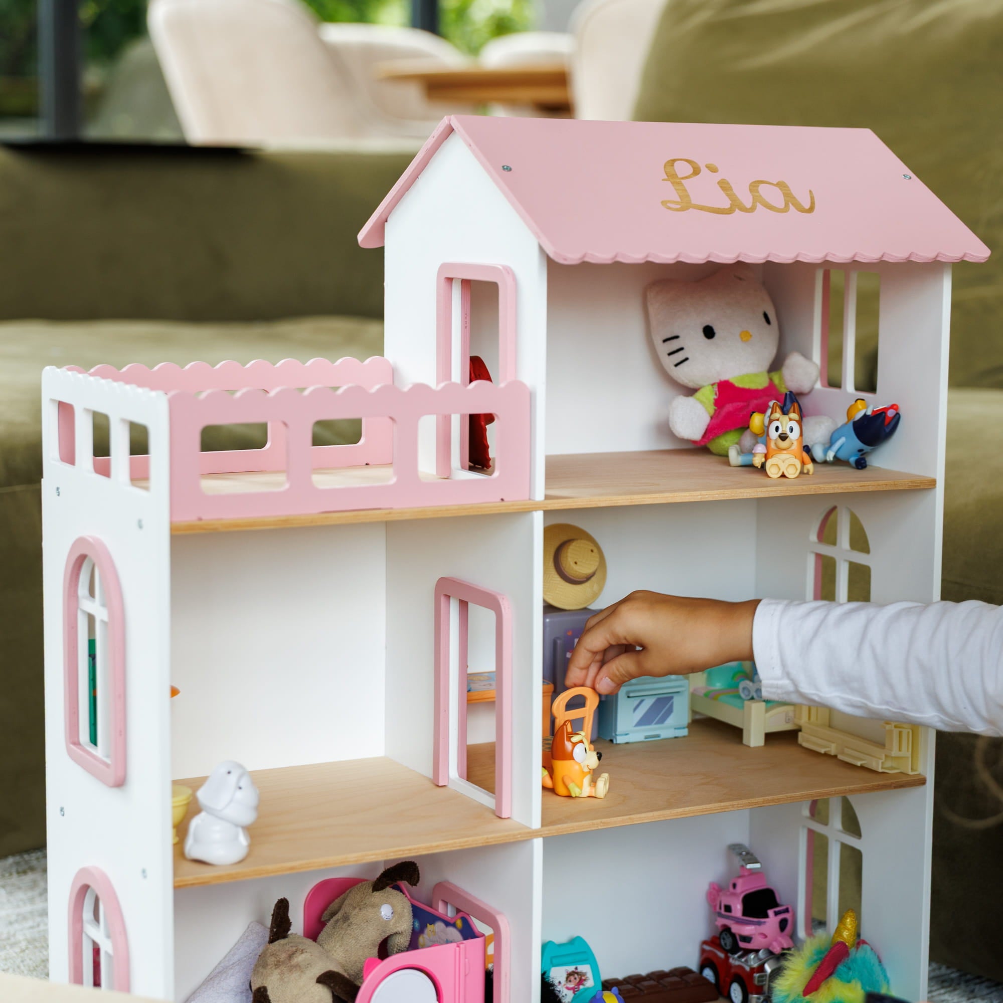 A white and pink wooden dollhouse with the name 'Lia' on the roof, shown in a living room setting with a child's hand placing a toy inside. The dollhouse is filled with various stuffed animals and toys.