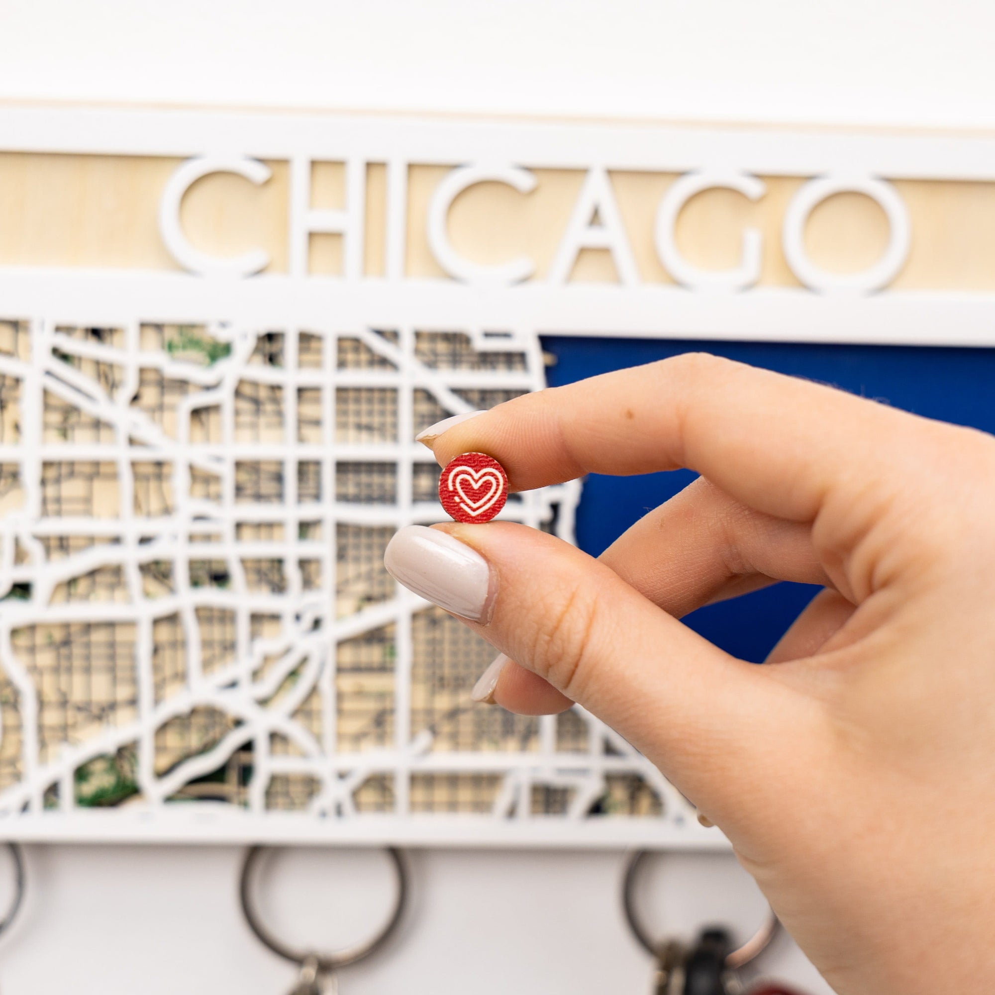 Hand holding a red heart-shaped keychain in front of a decorative sign with 'CHICAGO' on it.