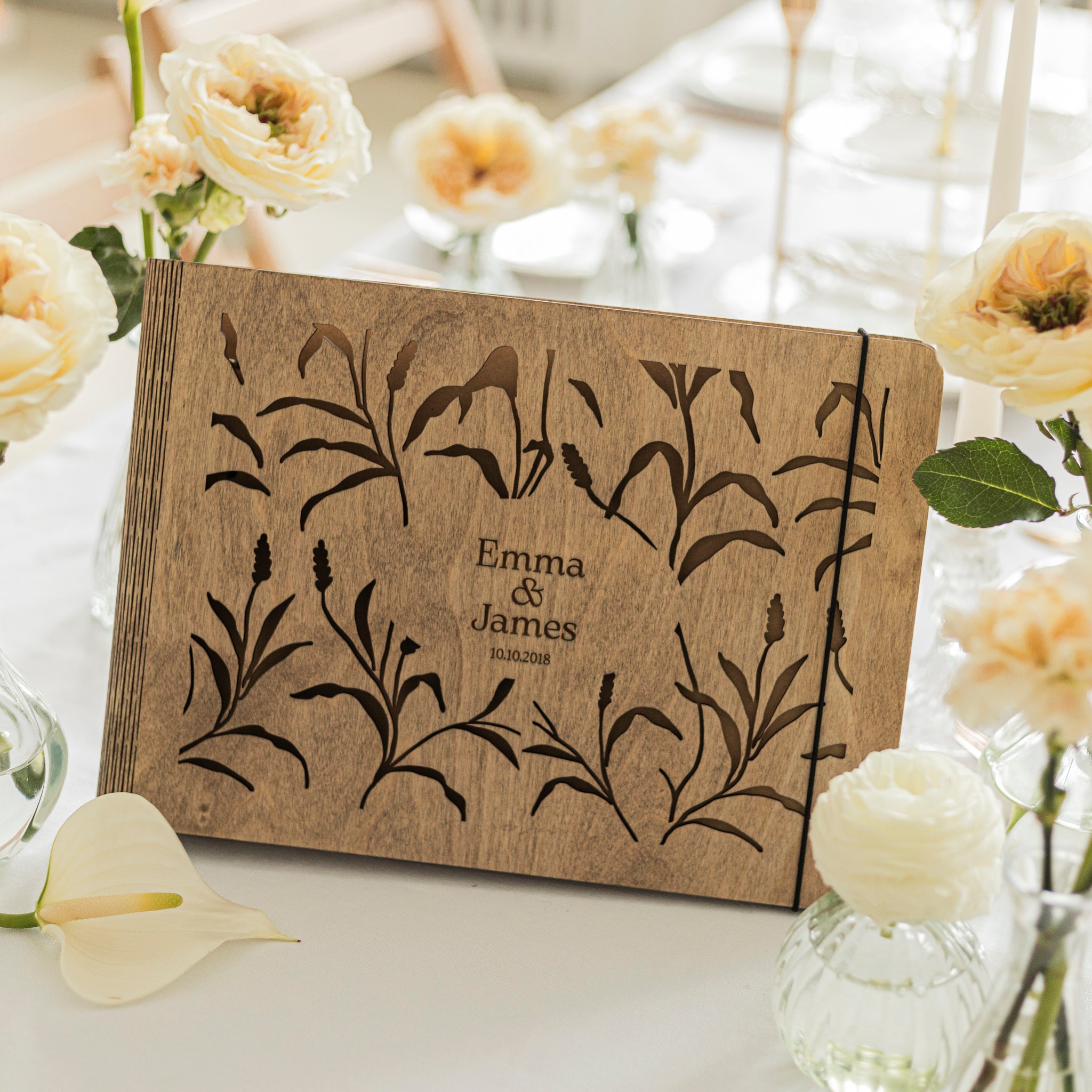 Wooden photo album with floral design and 'Emma & James' engraving, surrounded by white flowers.